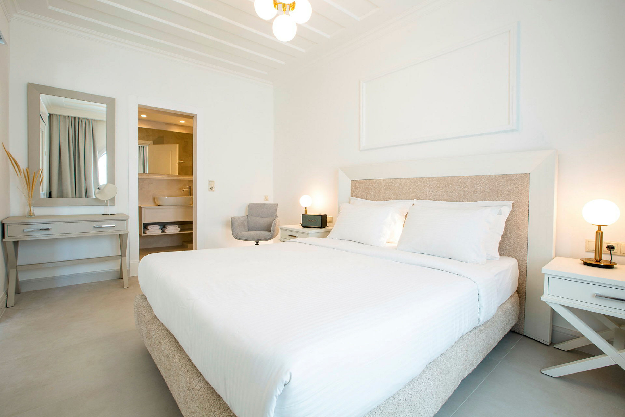 Et lyst og moderne hotellrom med en stor seng, to nattbord med lamper, et skrivebord med speil, og et åpent bad. (AI-generert tekst)