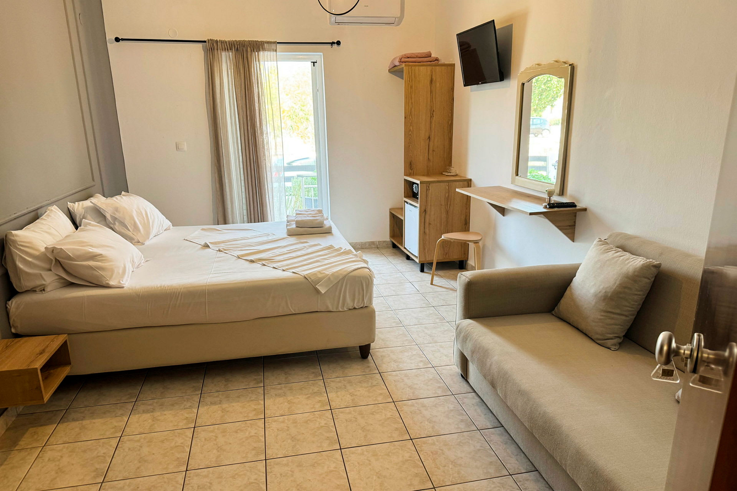 Et hotellrom med en seng, sofa, skrivebord og skap. Vinduet har lyse gardiner. (AI-generert tekst)