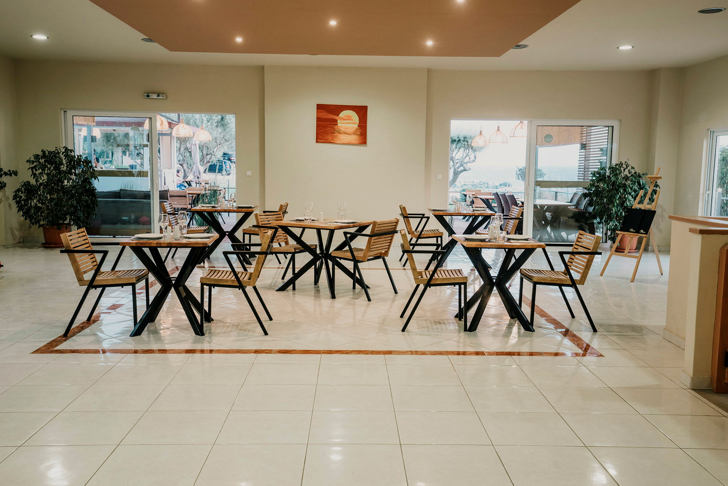 En restaurant med trestoler og bord, store vinduer og innendørsplante på gulvet. (AI-generert tekst)
