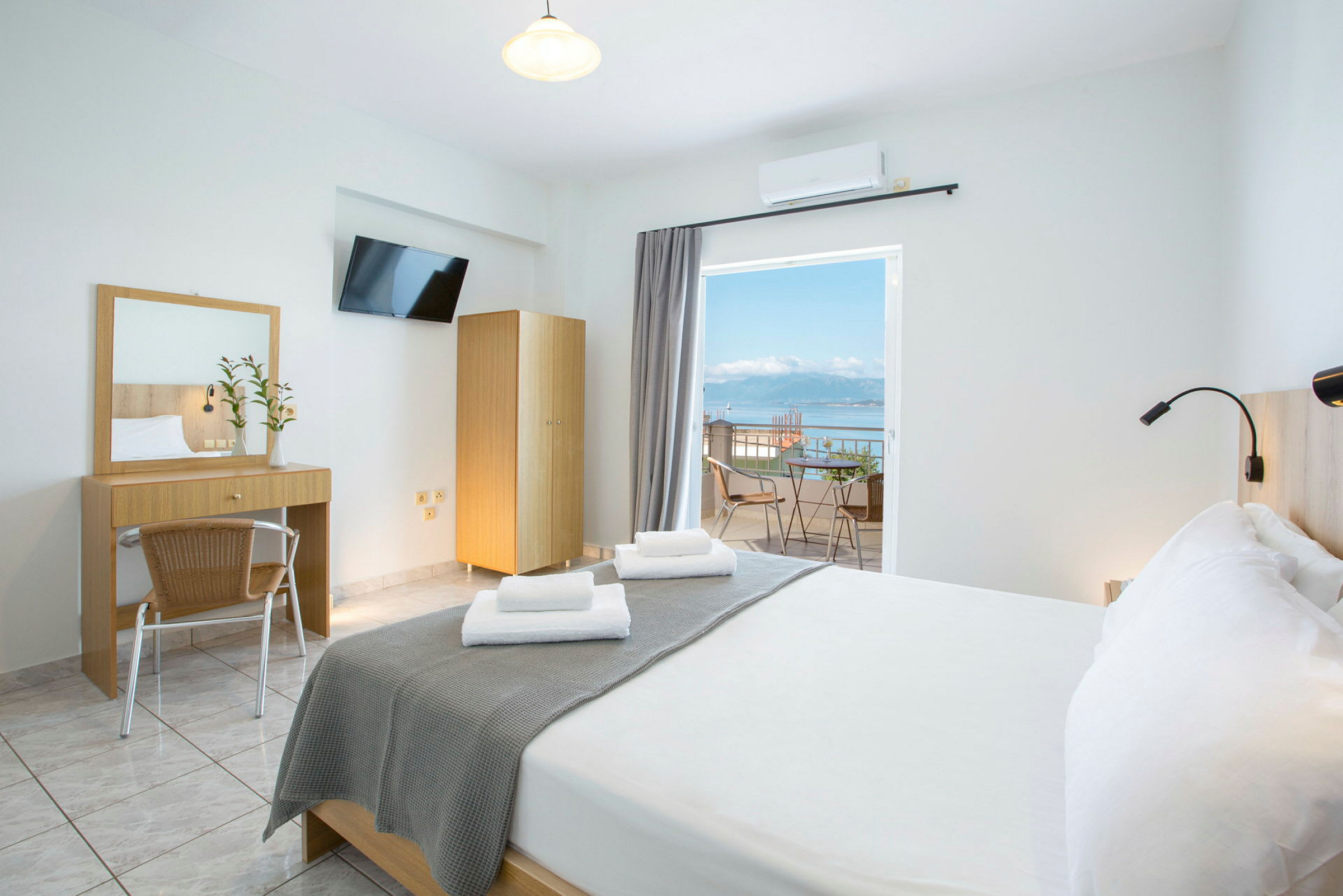 Et lyst hotellrom med en dobbeltseng, skrivebord, speil og en balkong med utsikt over havet. (AI-generert tekst)
