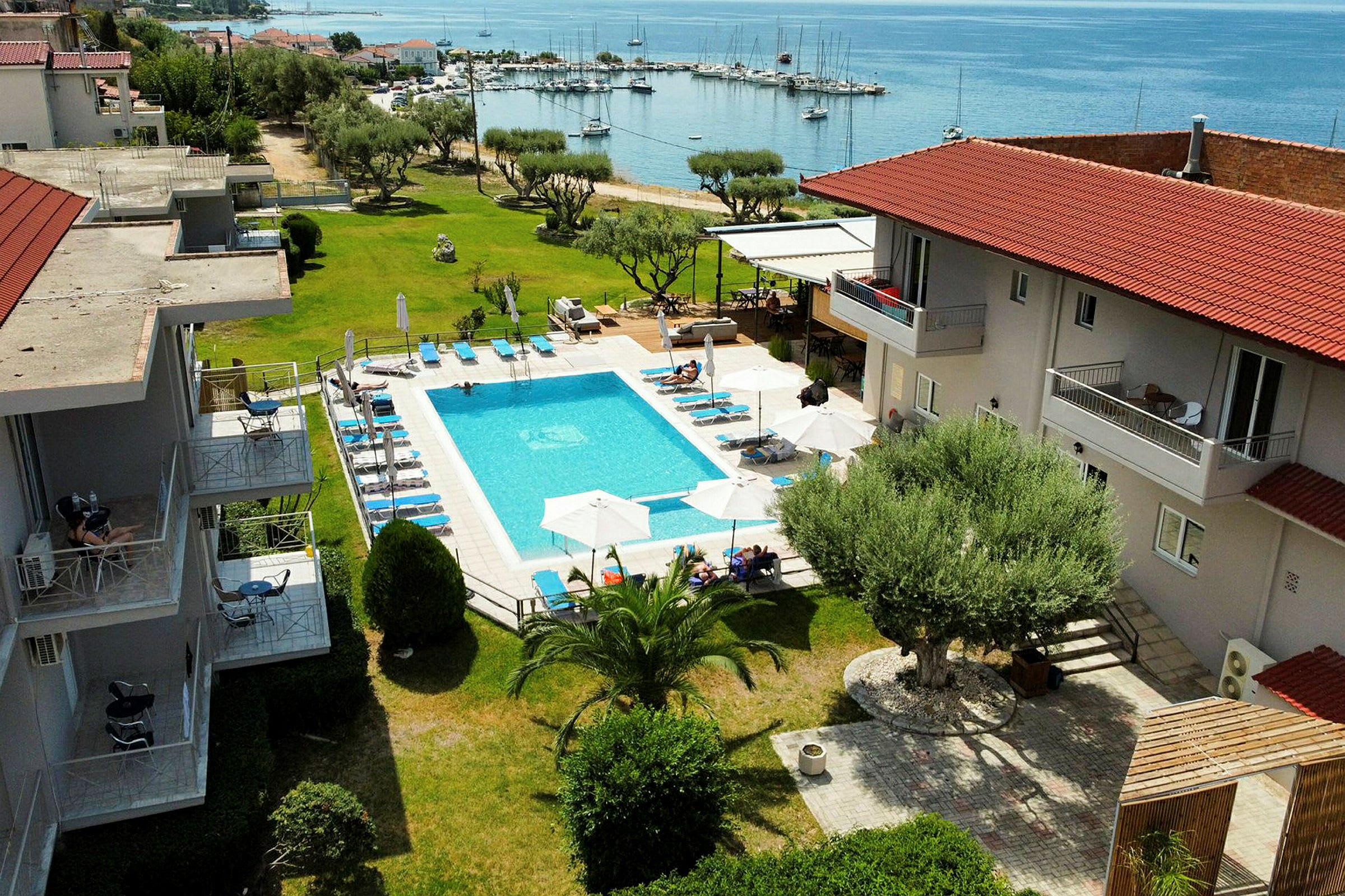 Bilde av et hotellområde med et utendørs basseng, solsenger og parasoller, samt omkringliggende grønne områder og utsikt mot en havn ved sjøen. (AI-generert tekst)