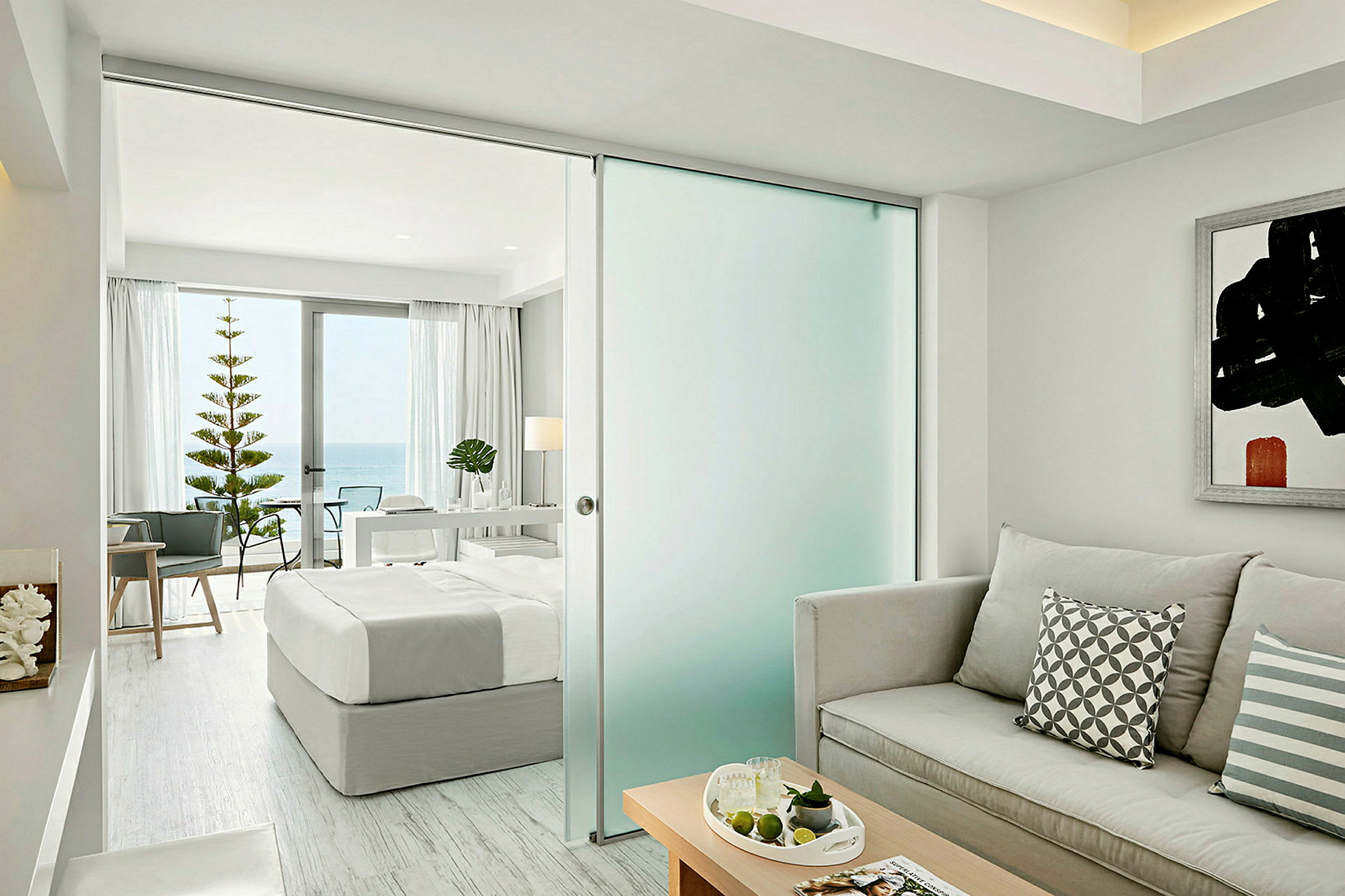 Et lyst og moderne hotellrom med en seng, sofa og utsikt mot havet gjennom store vinduer. (AI-generert tekst)
