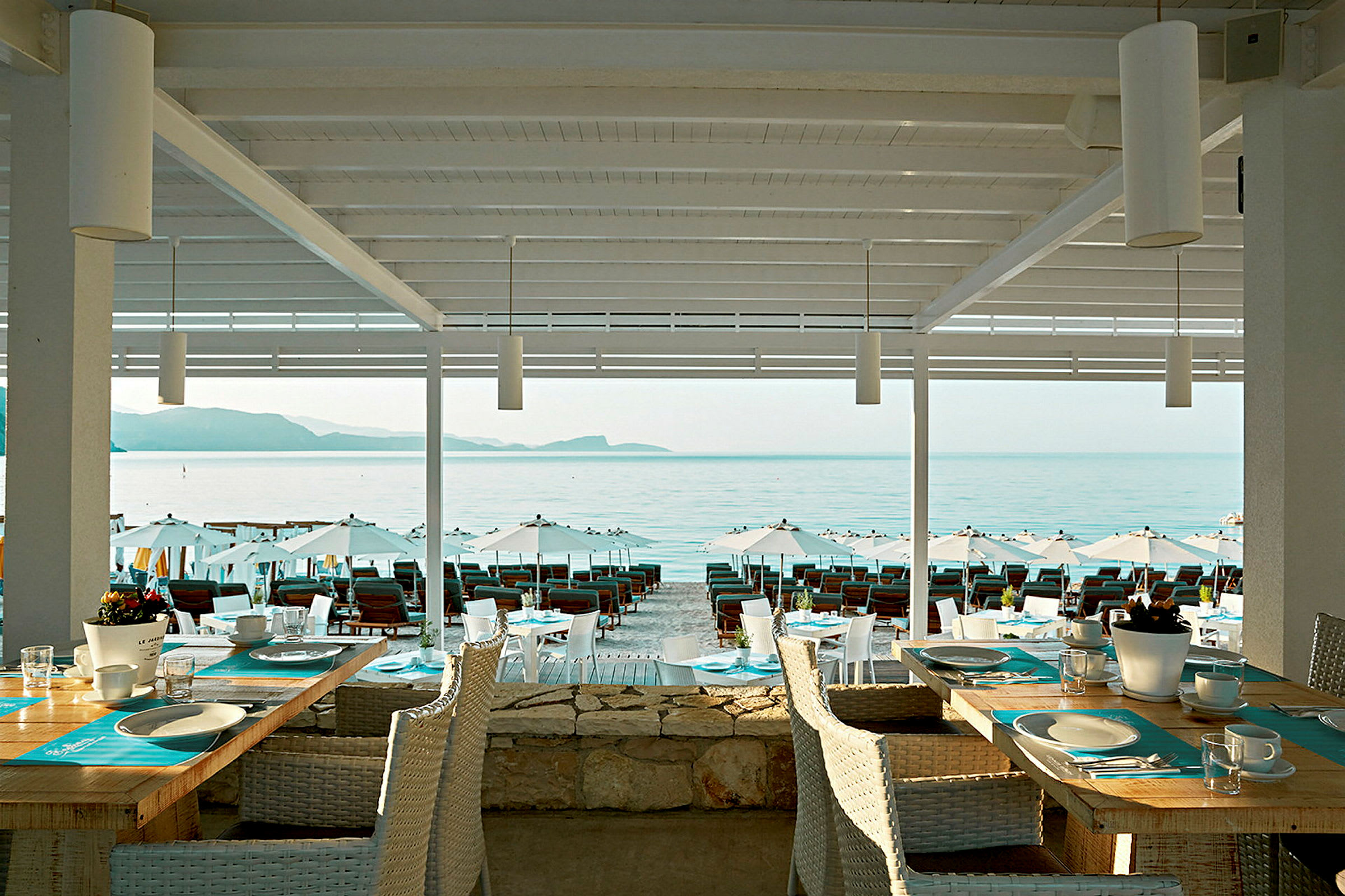 Utsikt fra en restaurant med dekkede bord mot stranden og havet. (AI-generert tekst)