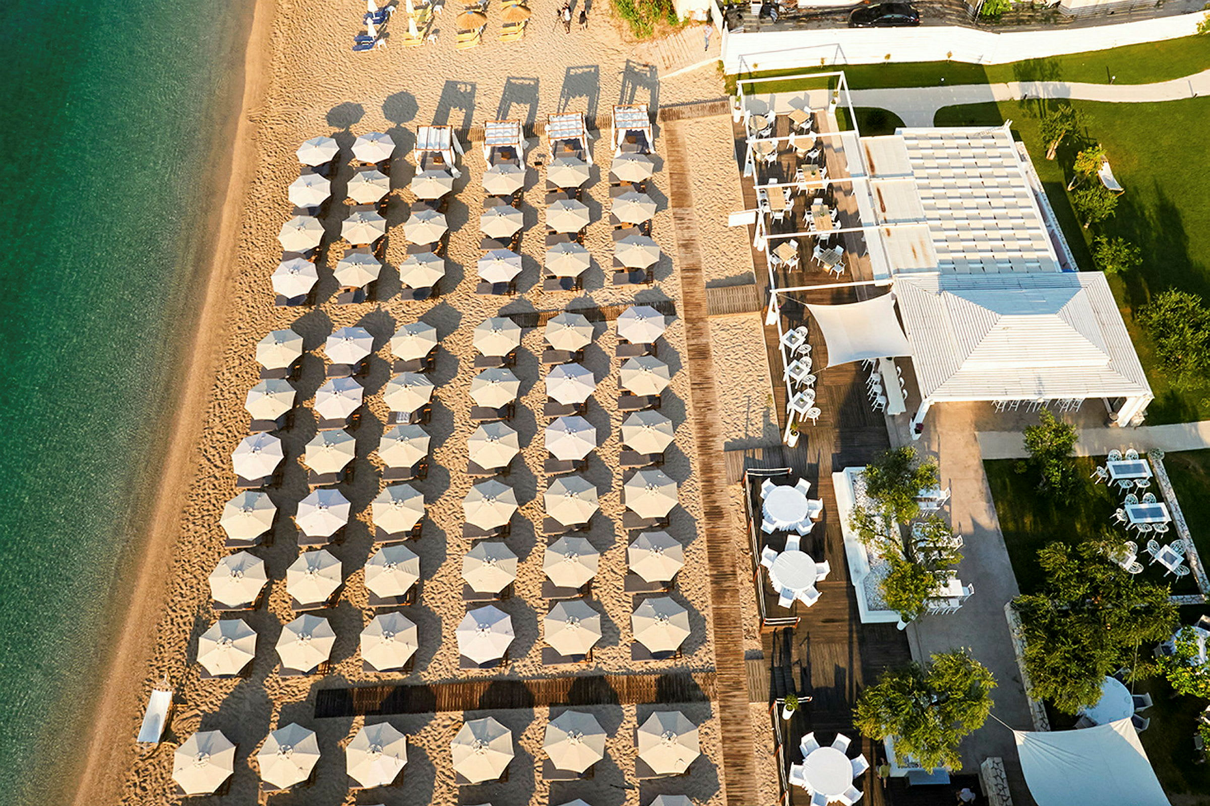 Flyfoto av en strand med oppstilte solsenger og parasoller samt en restaurant med sitteplasser. (AI-generert tekst)