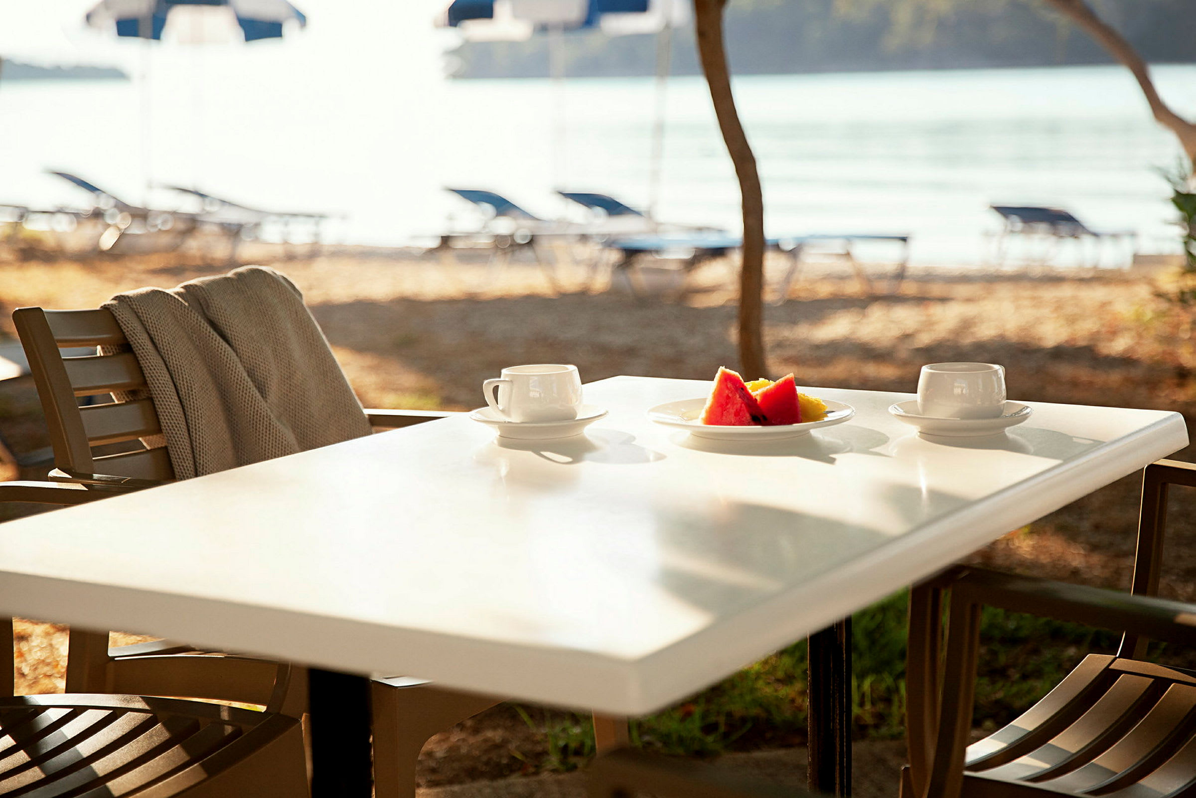 En utendørs servering med et bord med to kaffekopper og en tallerken med frukt. Bakgrunnen viser strand og vann. (AI-generert tekst)