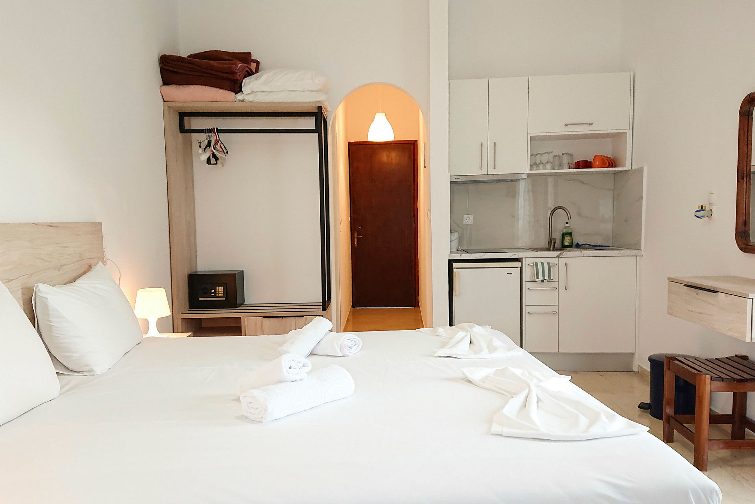 Hotellrom med en oppredd dobbeltseng, garderobe, dør, kjøkkenkrok og stol. (AI-generert tekst)