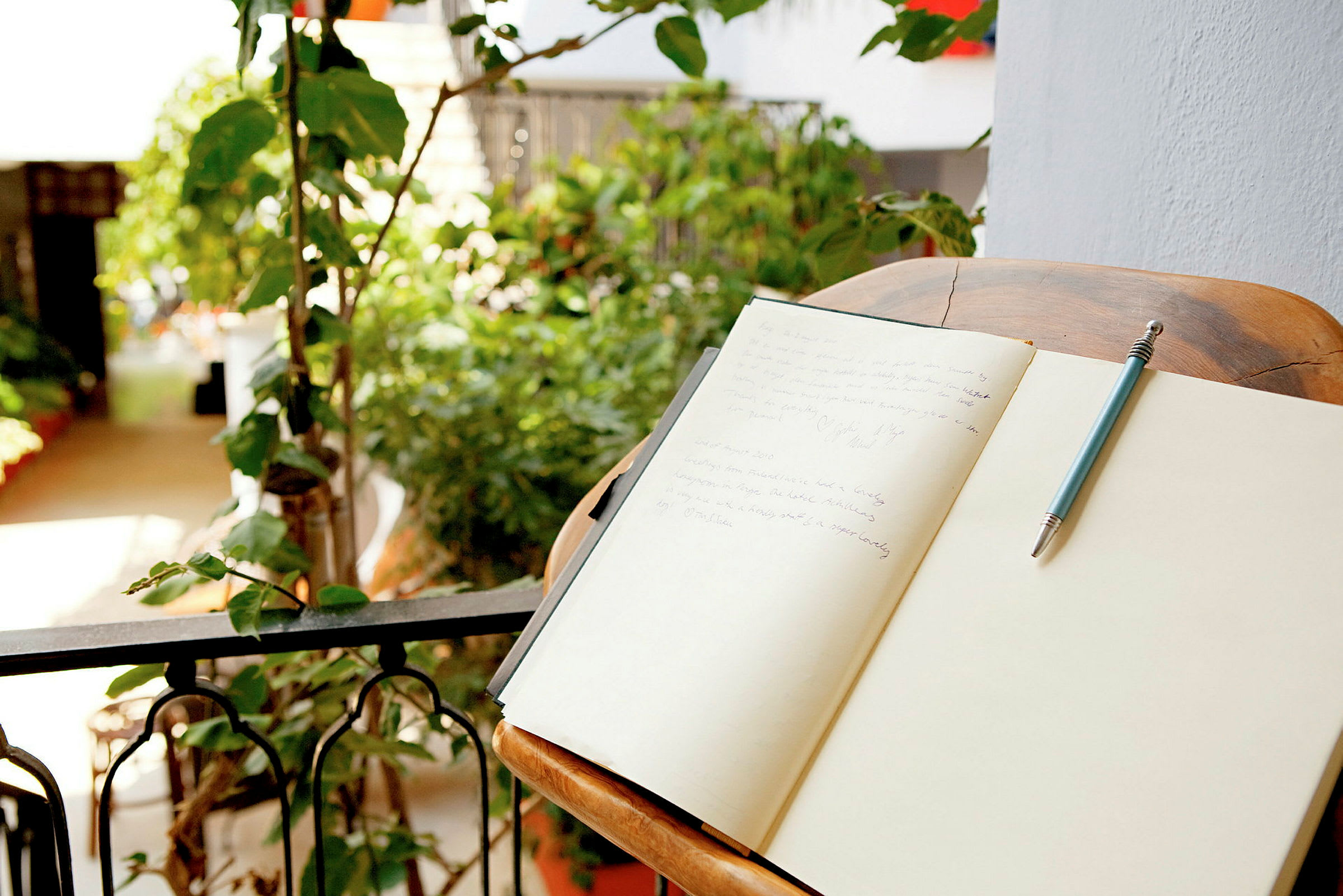 Et skrivebord med en åpen notatbok og en penn ved siden av, plassert på en balkong med planter i bakgrunnen. (AI-generert tekst)