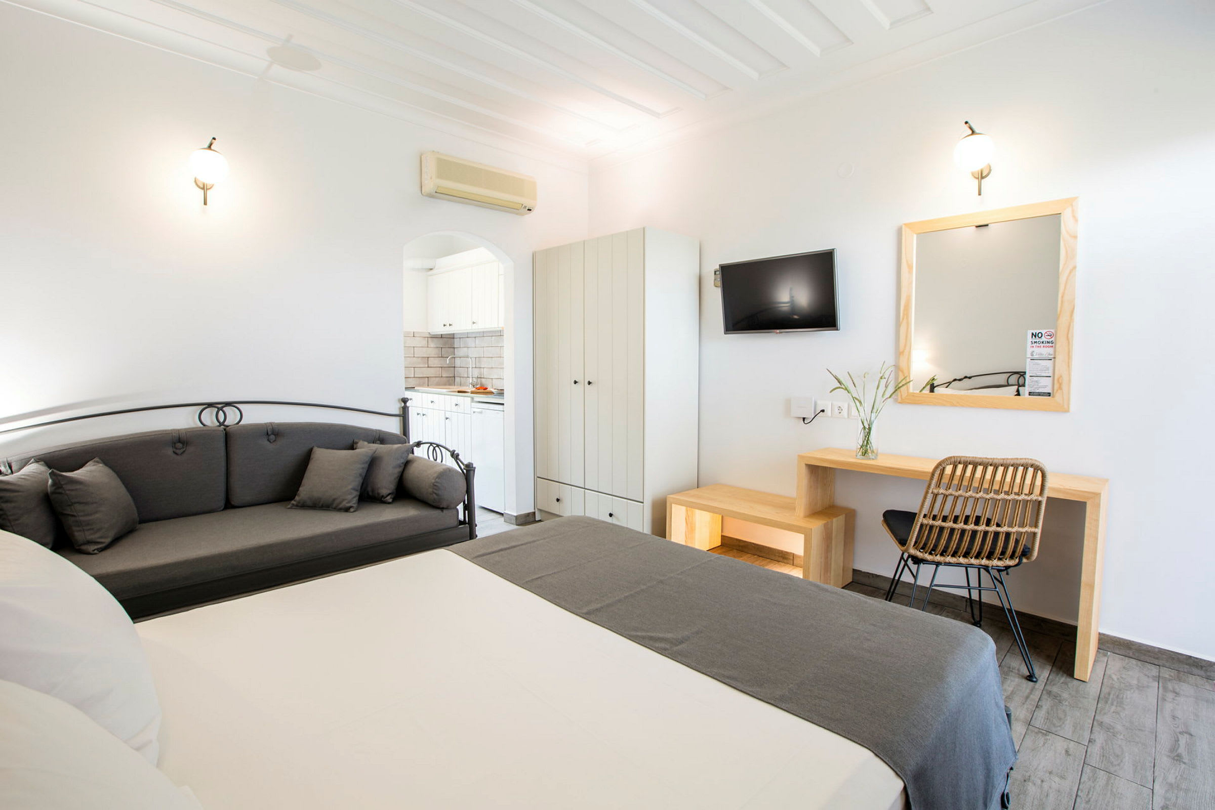 Et hotellrom med seng, grå sofa, skrivebord, veggmontert TV og garderobe. Et bad vises gjennom en døråpning. (AI-generert tekst)