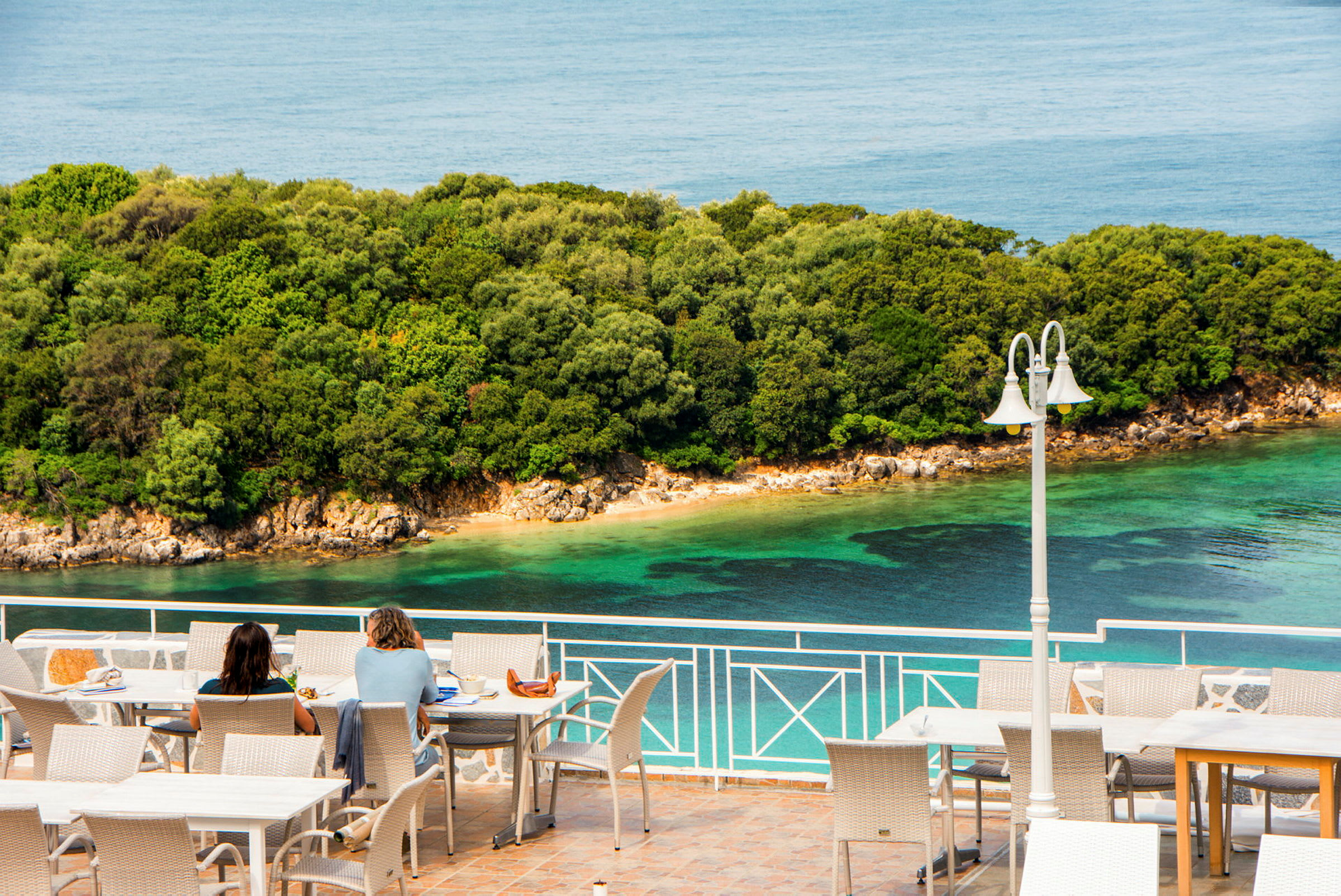 To personer sitter ved bord på en terrasse med utsikt over havet og en frodig kystlinje. (AI-generert tekst)