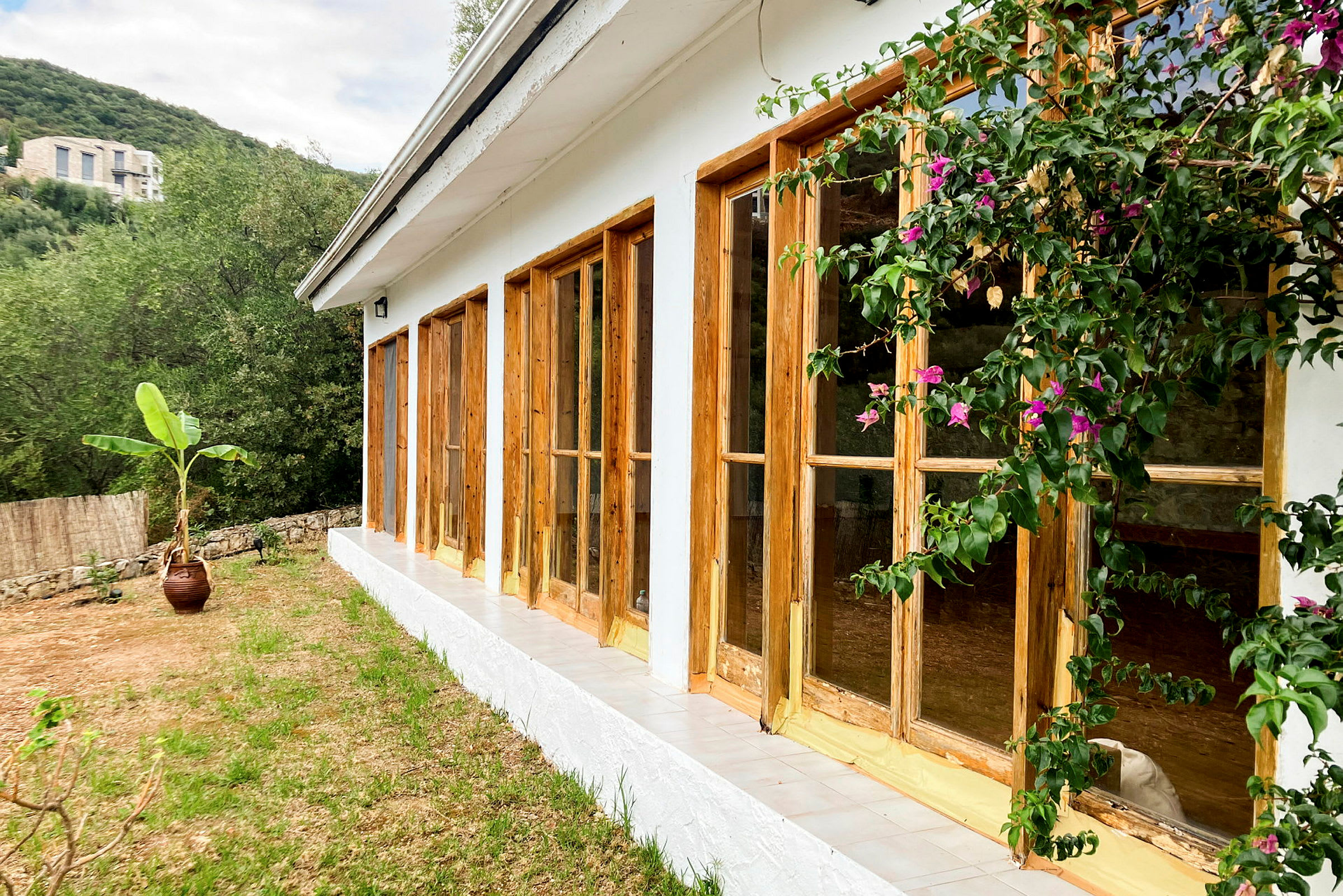 Bilde av et hus med hvite vegger og vinduer med trerammer, omgitt av grøntområder og blomster. (AI-generert tekst)