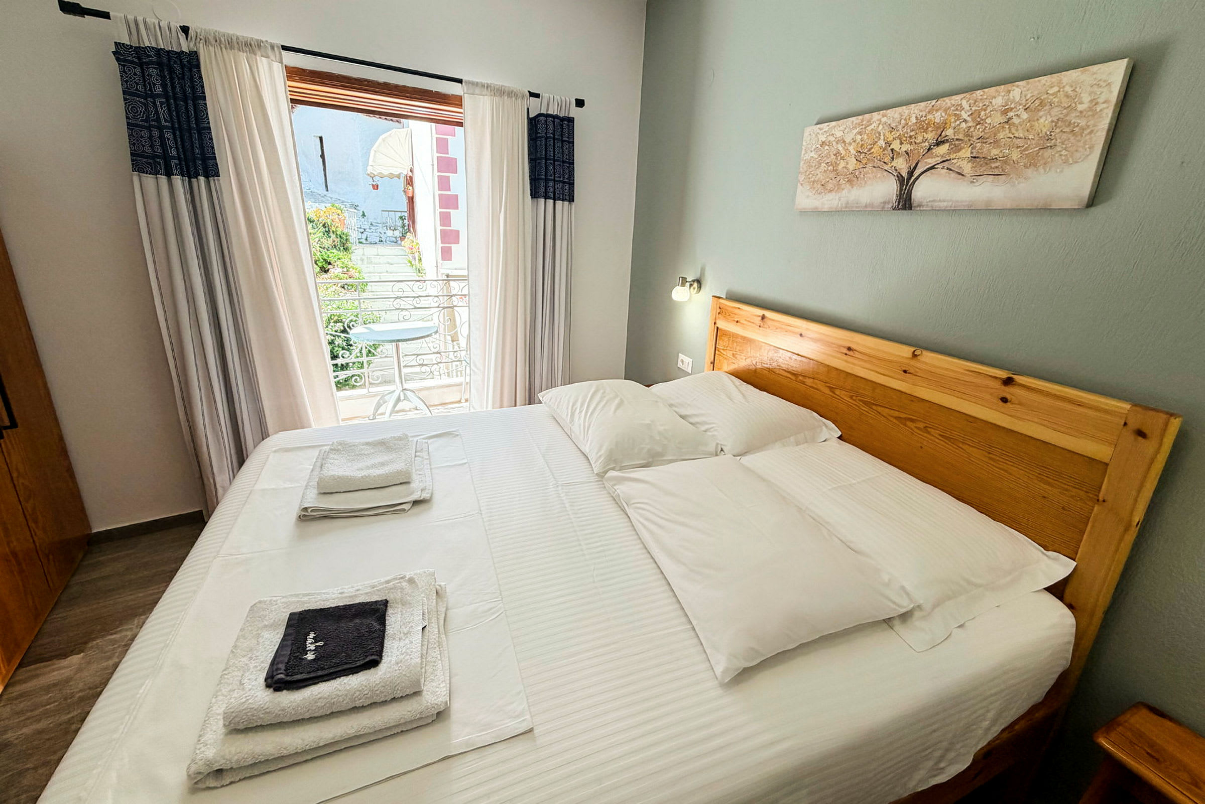Hotellrom med dobbeltseng, hvitt sengetøy og tredesign på sengegavlen. Balkongutsikt kan ses gjennom glassdørene. (AI-generert tekst)