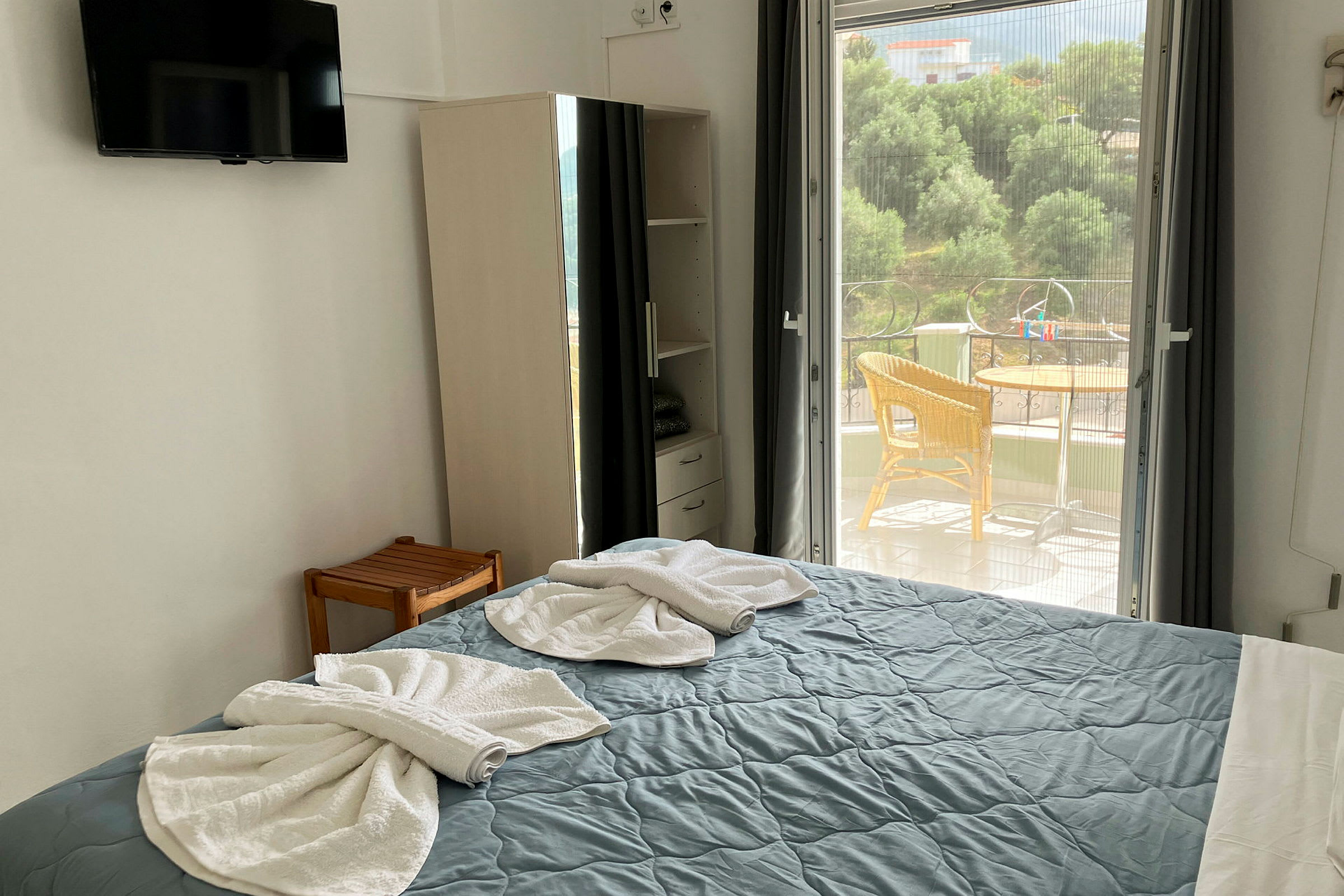 Et hotellrom viser en oppredd seng med foldede håndklær, et skap og en utgang til en balkong. (AI-generert tekst)