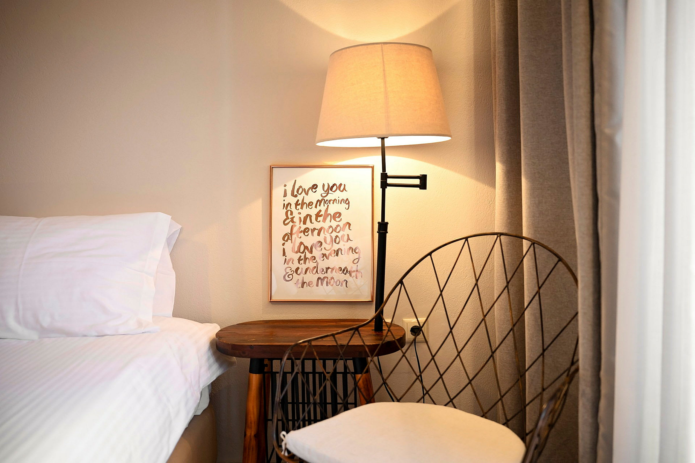 Et nattbord med en lampe og en stol ved siden av en seng. En tavle med tekst henger på veggen. (AI-generert tekst)
