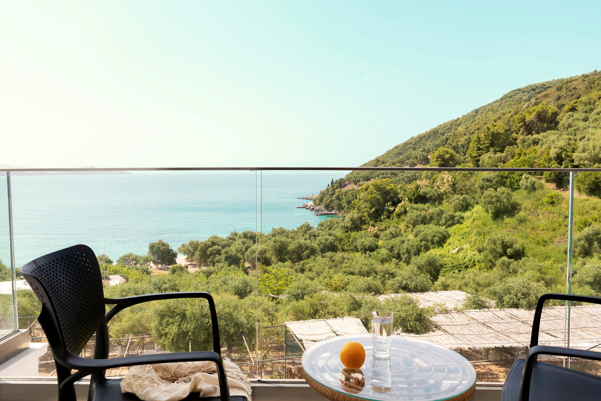 Utsikt fra en terrasse med stoler og et bord mot havet og grønne skogkledde åser. (AI-generert tekst)