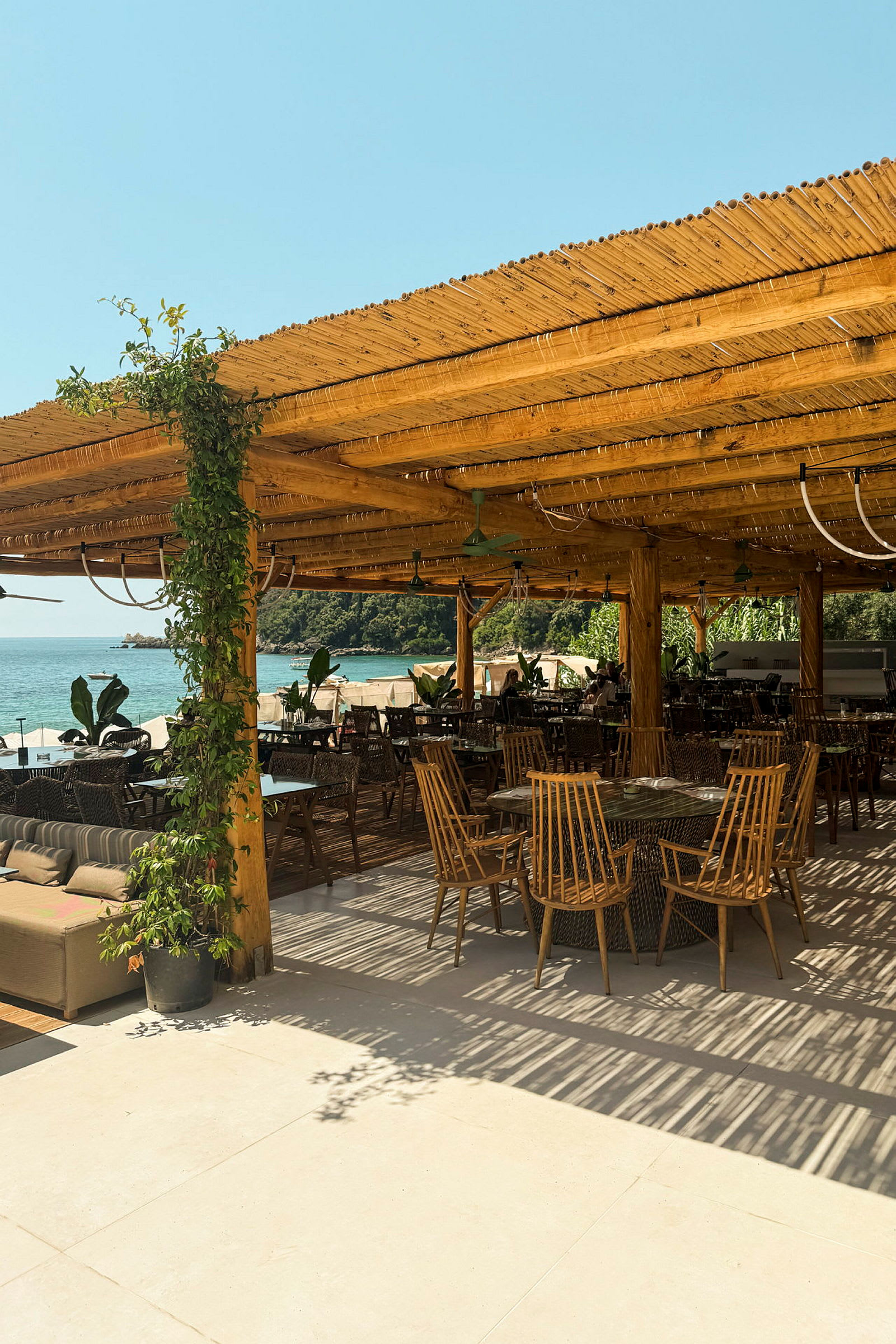 Utendørs servering med trepergola, bord og stoler samt utsikt mot havet. (AI-generert tekst)