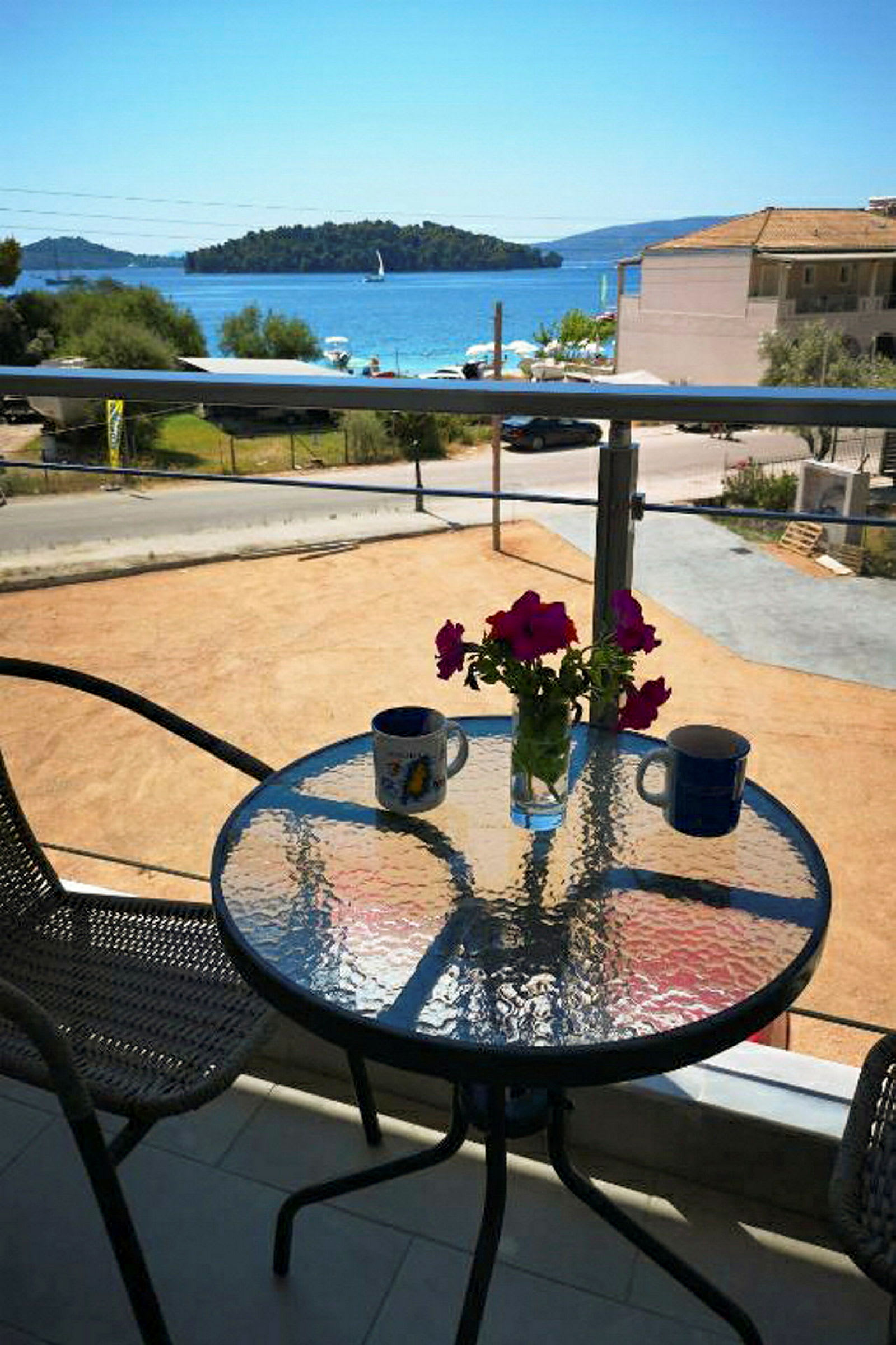 En balkong med et glassbord, to kopper og en vase med rosa blomster, med utsikt over havet og en liten øy. (AI-generert tekst)