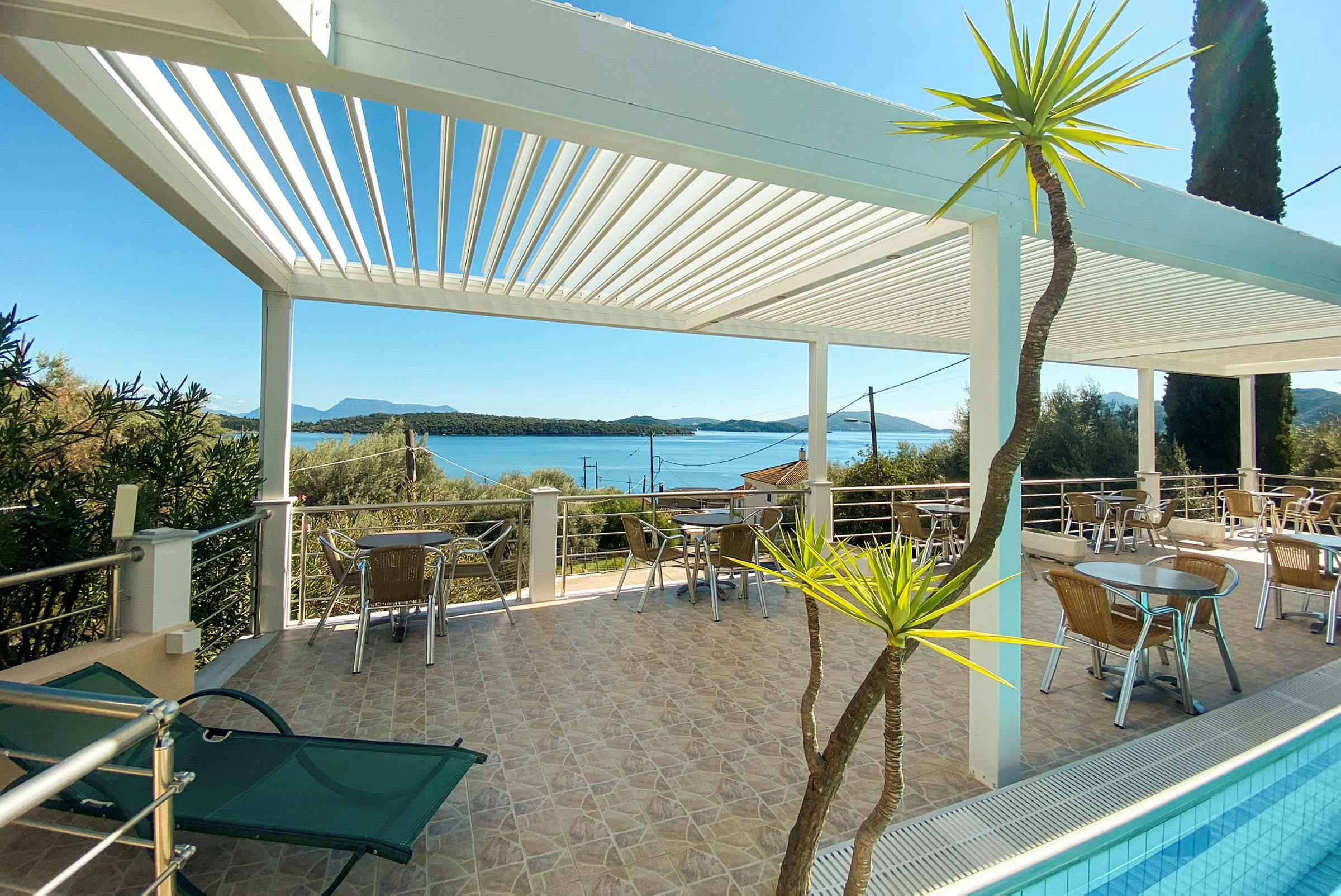Terrasse med sitteplasser under en hvit pergola, utsikt over havet og omkringliggende vegetasjon. (AI-generert tekst)