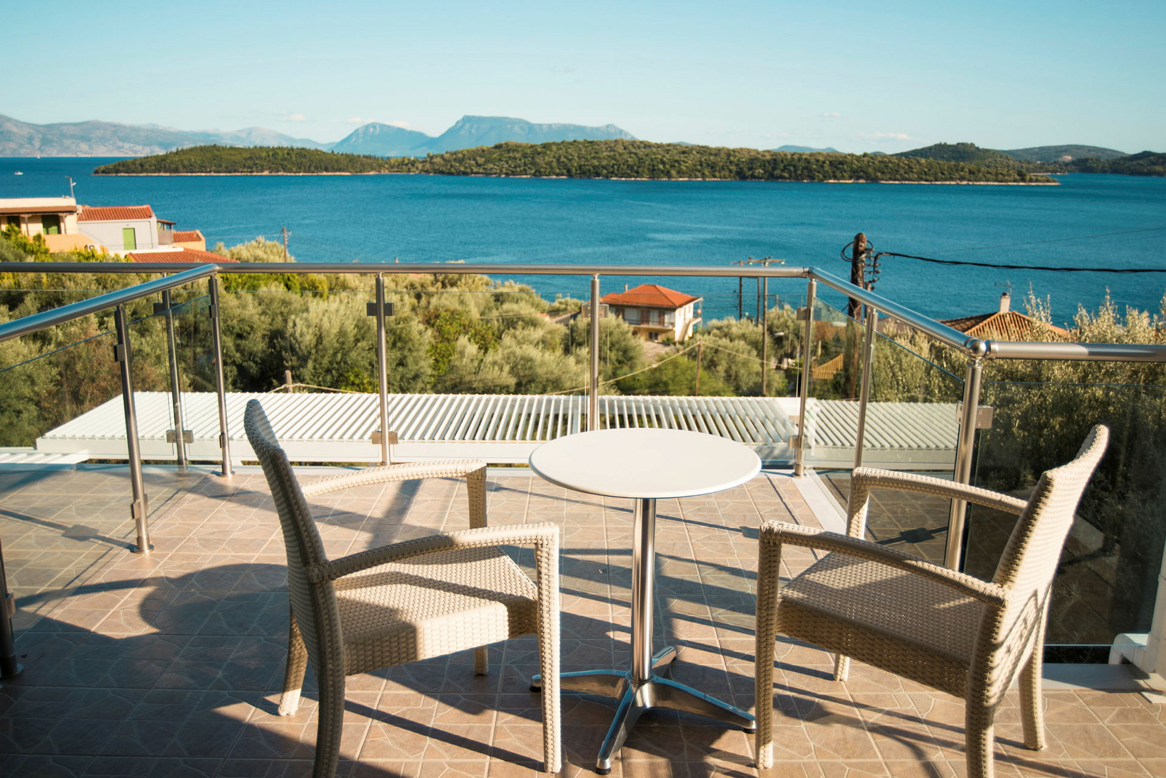 To stoler og et lite bord på en terrasse med glassrekkverk, utsikt mot havet og skogkledde øyer. (AI-generert tekst)