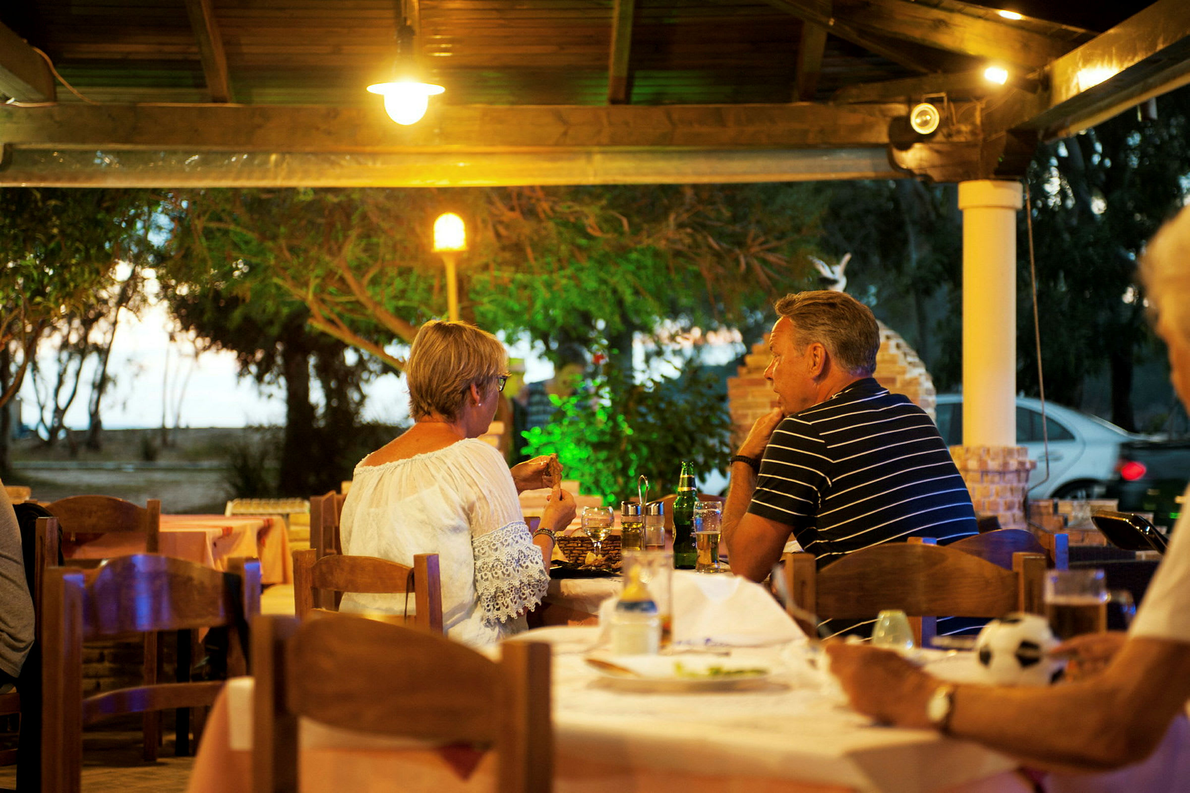 Personer sitter ved et bord på en utendørsrestaurant omgitt av grøntområder. (AI-generert tekst)