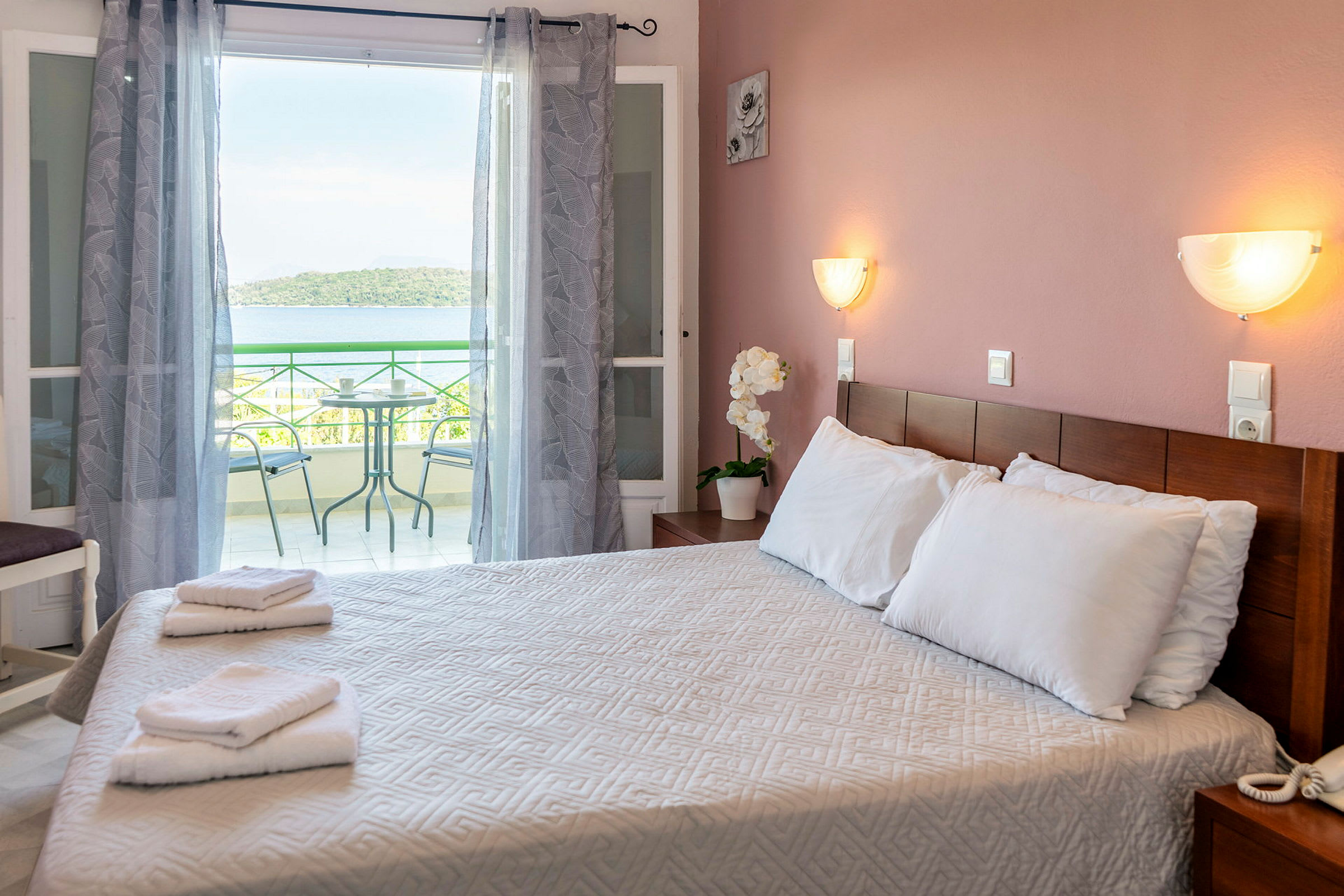 Et hotellrom med en dobbeltseng, håndklær på sengen og en balkong i bakgrunnen med utsikt mot havet. (AI-generert tekst)