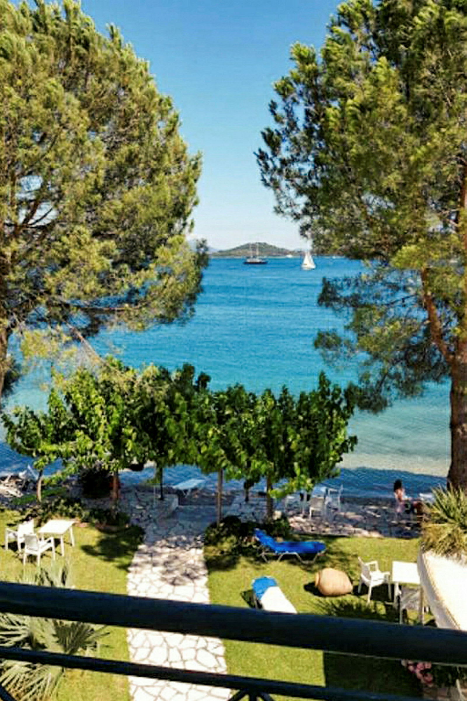 Utsikt fra en terrasse med trær, plen, stoler og bord, med havet i bakgrunnen. (AI-generert tekst)