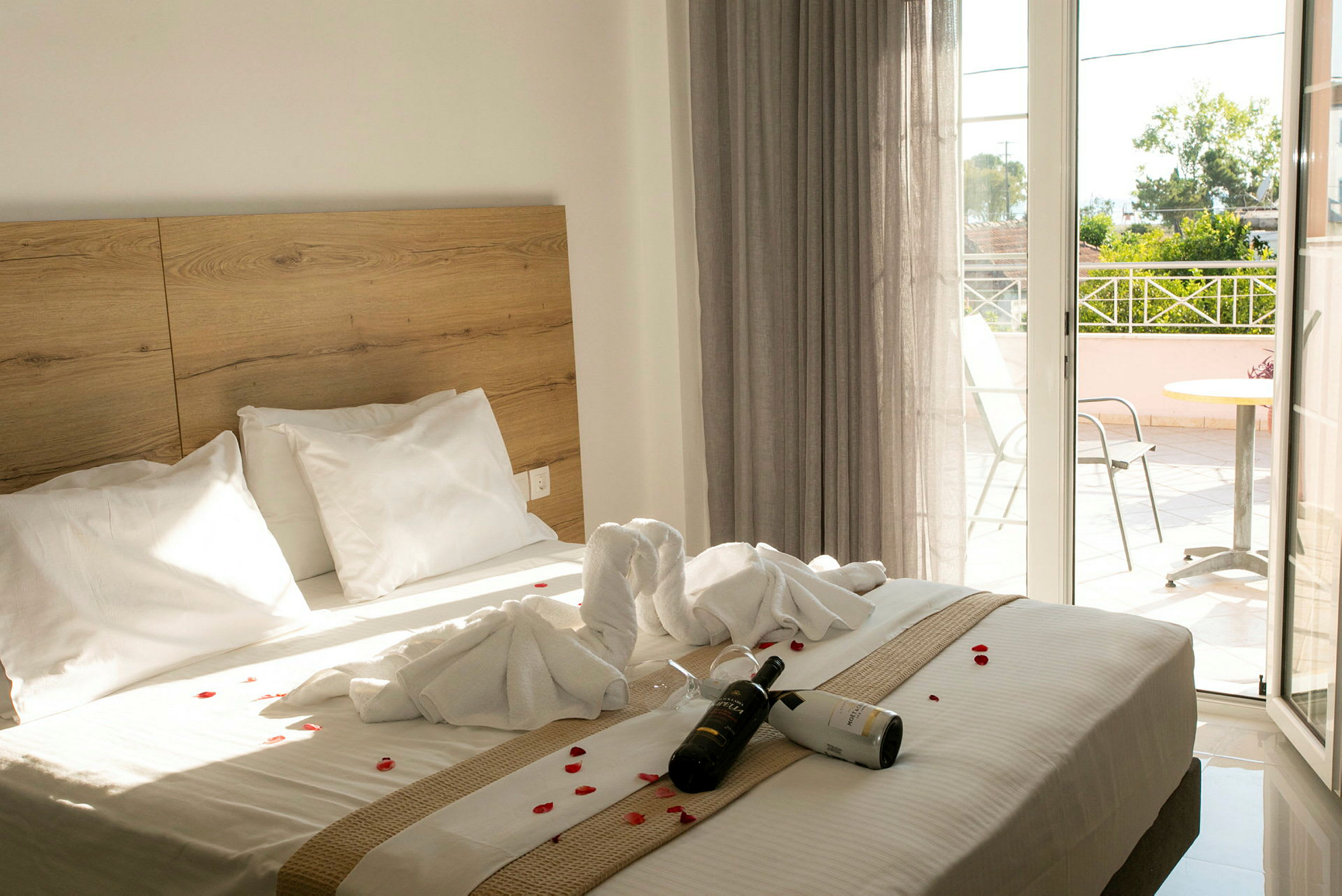 Hotellrom med en dobbeltseng dekorert med håndklær og blomsterblader. En flaske vin og glass ligger på sengen. En balkong er synlig i bakgrunnen. (AI-generert tekst)