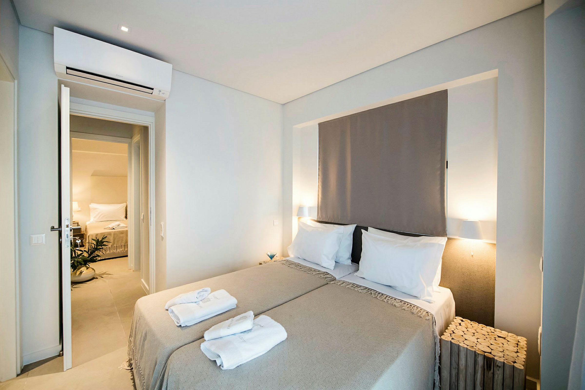 Et soverom med to enkeltsenger dekket med beige sengetøy og sammenbrettede håndklær plassert oppå. En dør åpnes mot et annet rom med en seng og en potteplante. (AI-generert tekst)