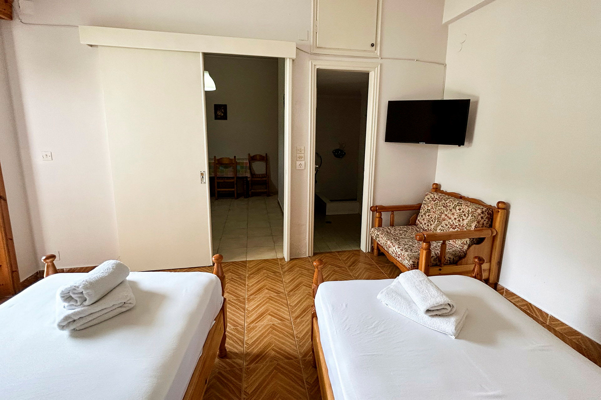 Et hotellrom med to senger, en lukket garderobe, en liten sittemøbel og en TV på veggen. (AI-generert tekst)