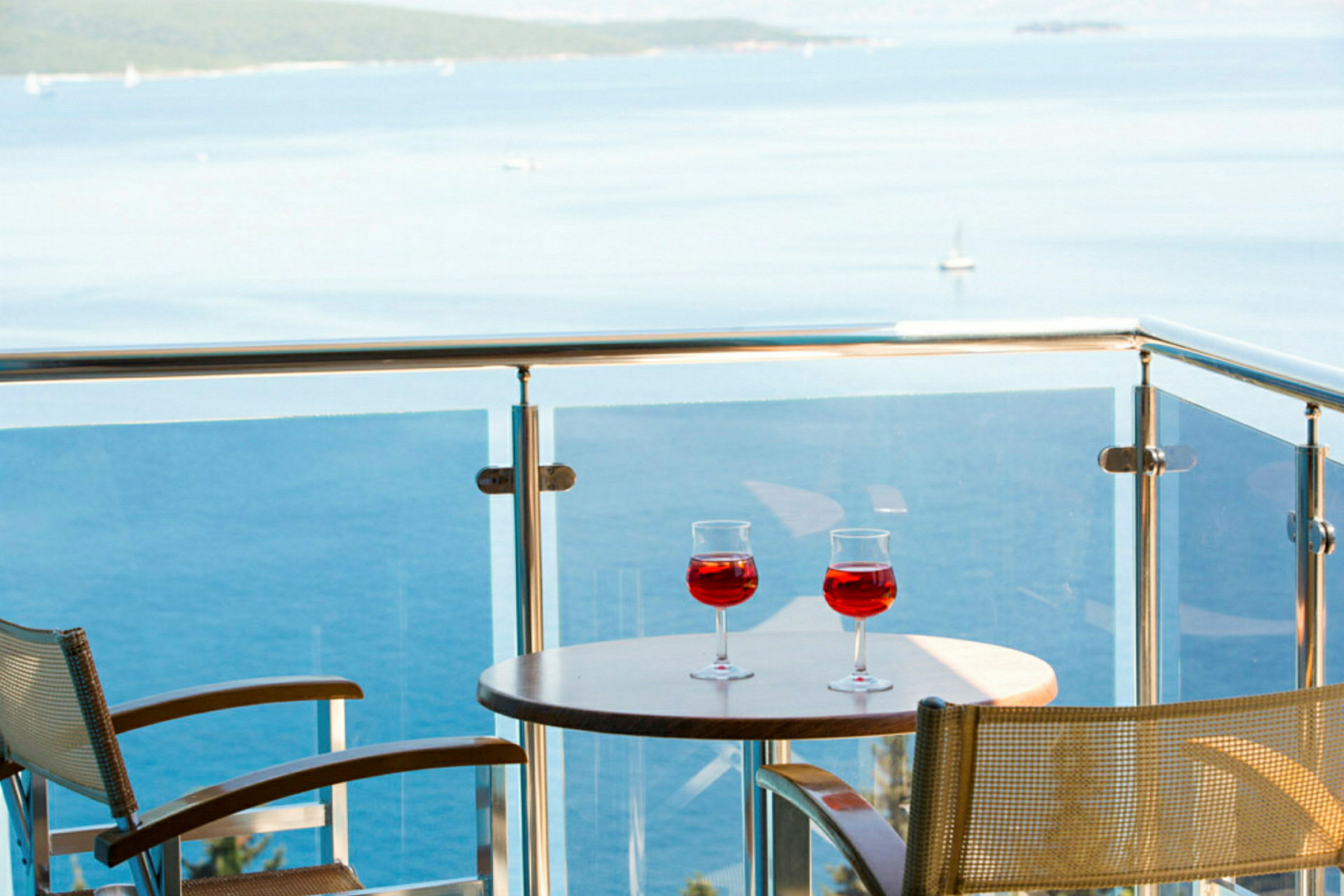 To glass med rød drikk på et bord på en balkong med utsikt over havet. (AI-generert tekst)