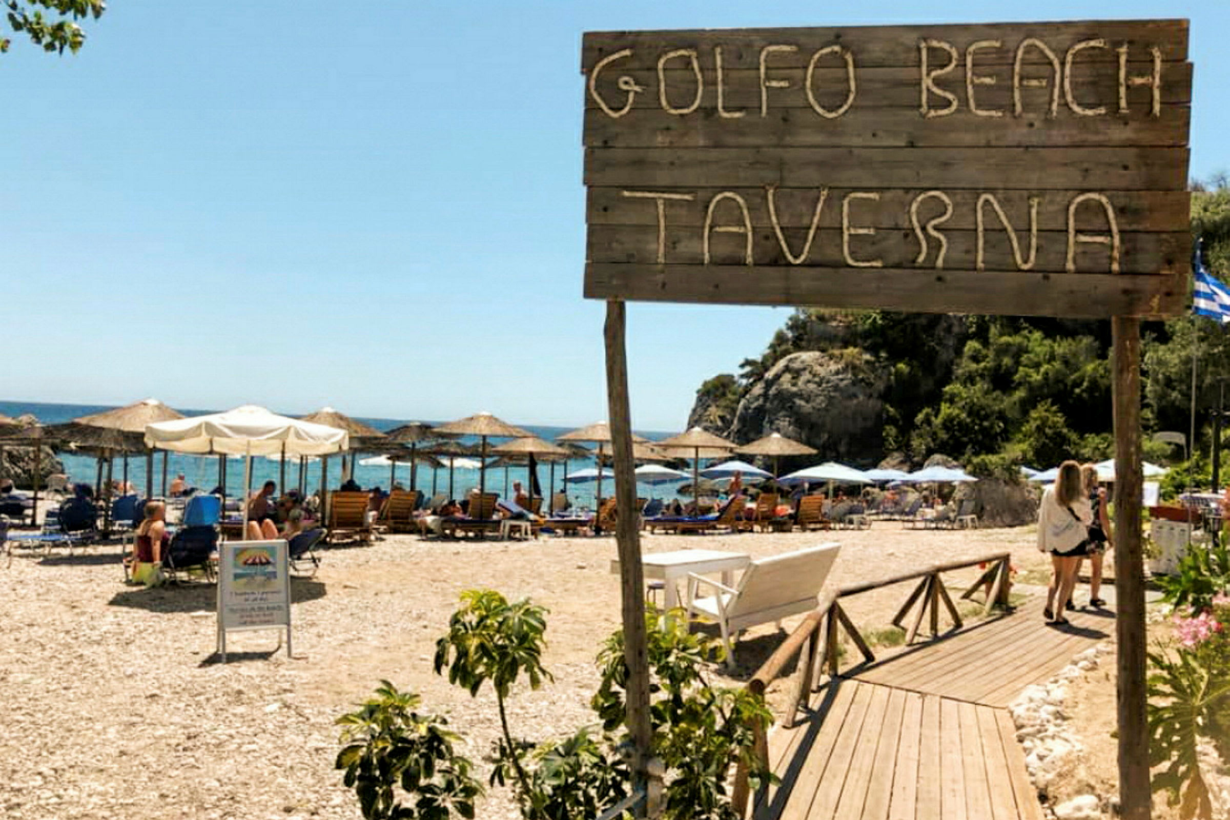 Skilt med teksten 'Golfo Beach Taverna' foran en strand med parasoller og solsenger. (AI-generert tekst)
