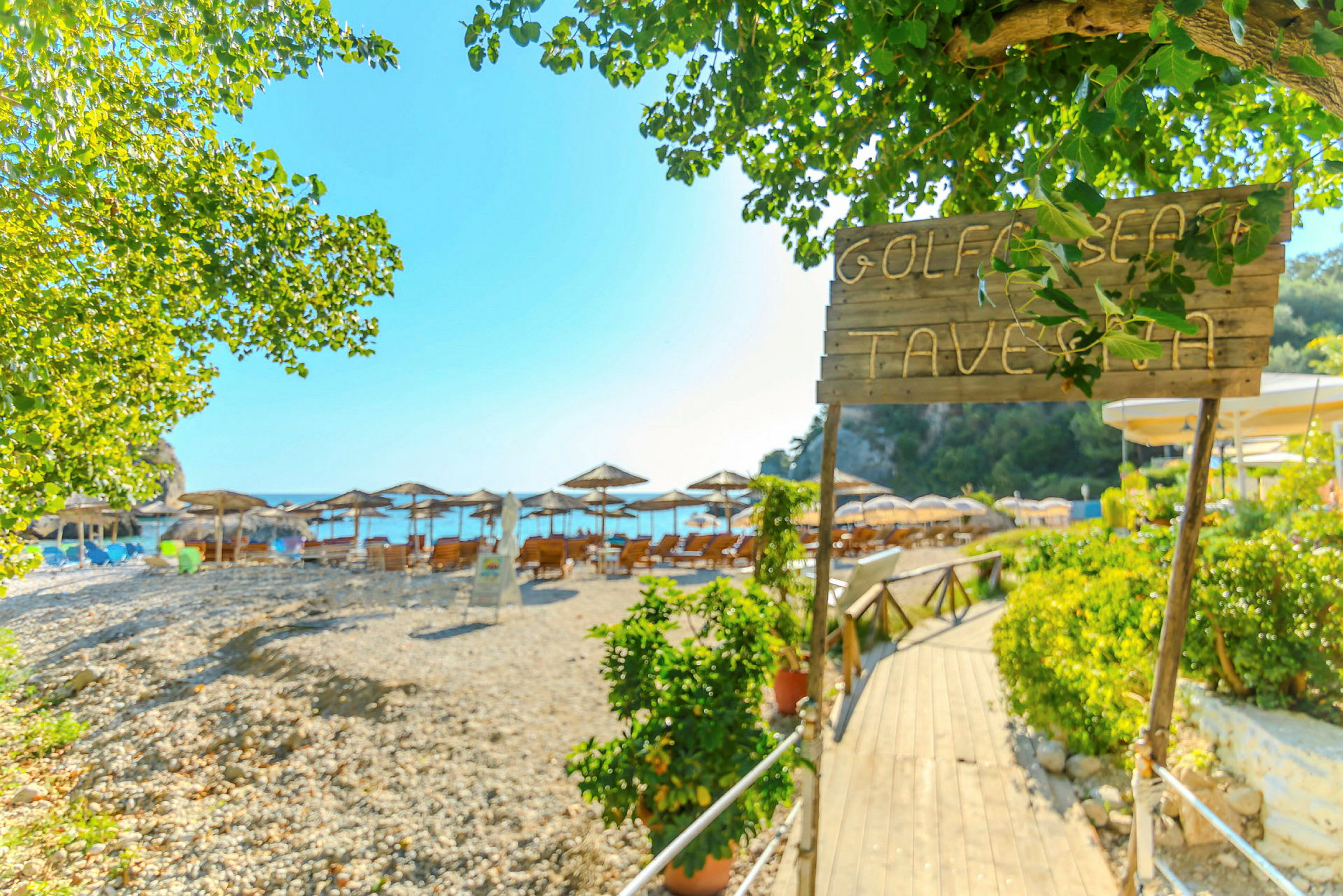 Skilt med teksten 'Golf Beach Taverna' ved inngangen til en strand med parasoller og solsenger. (AI-generert tekst)