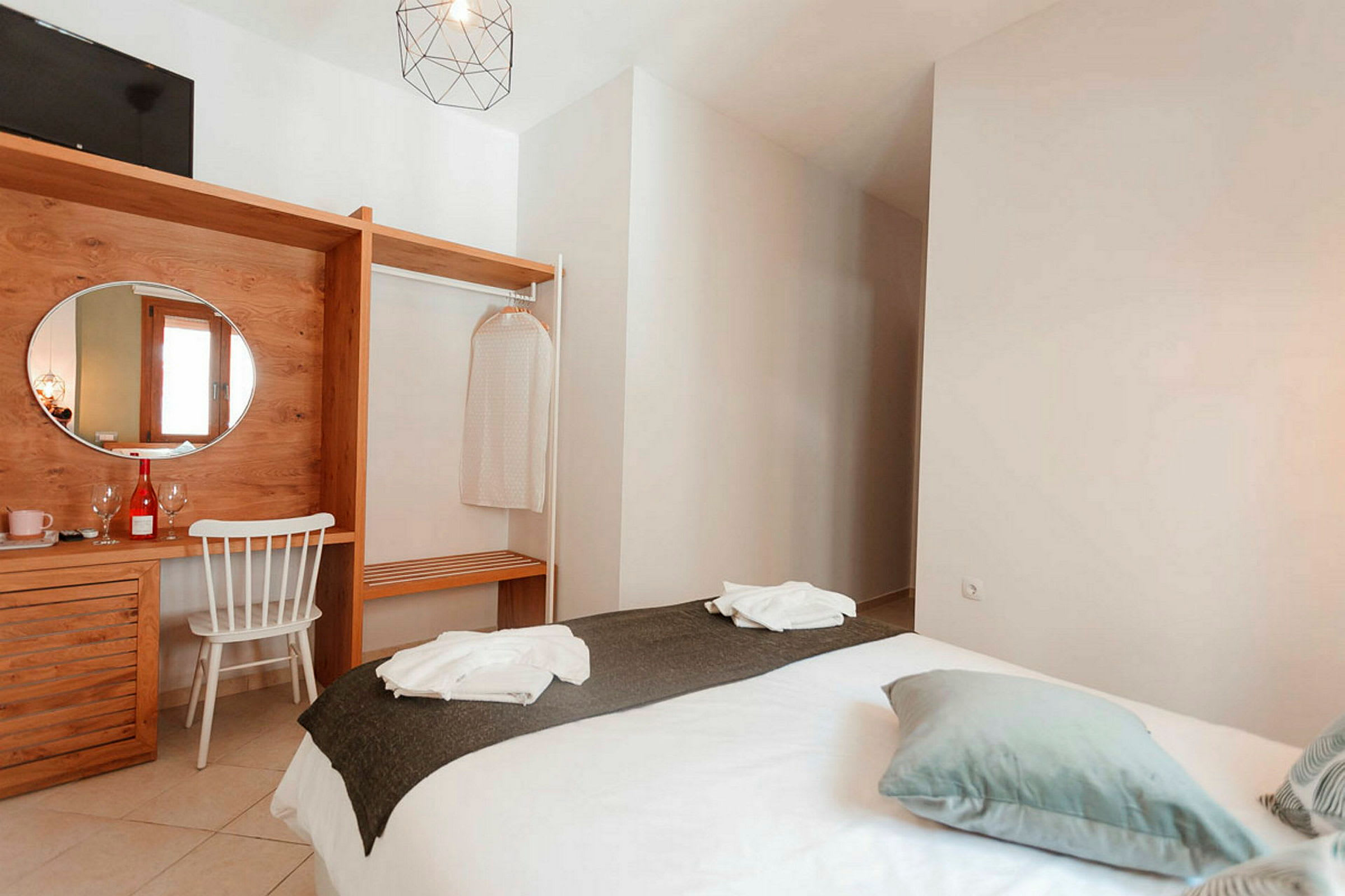 Et hotellrom med en seng kledd i hvitt sengetøy og mørkegrått overkast, en treinnredning med et speil og en stol, samt detaljer som kleshengere og dekorasjon. (AI-generert tekst)