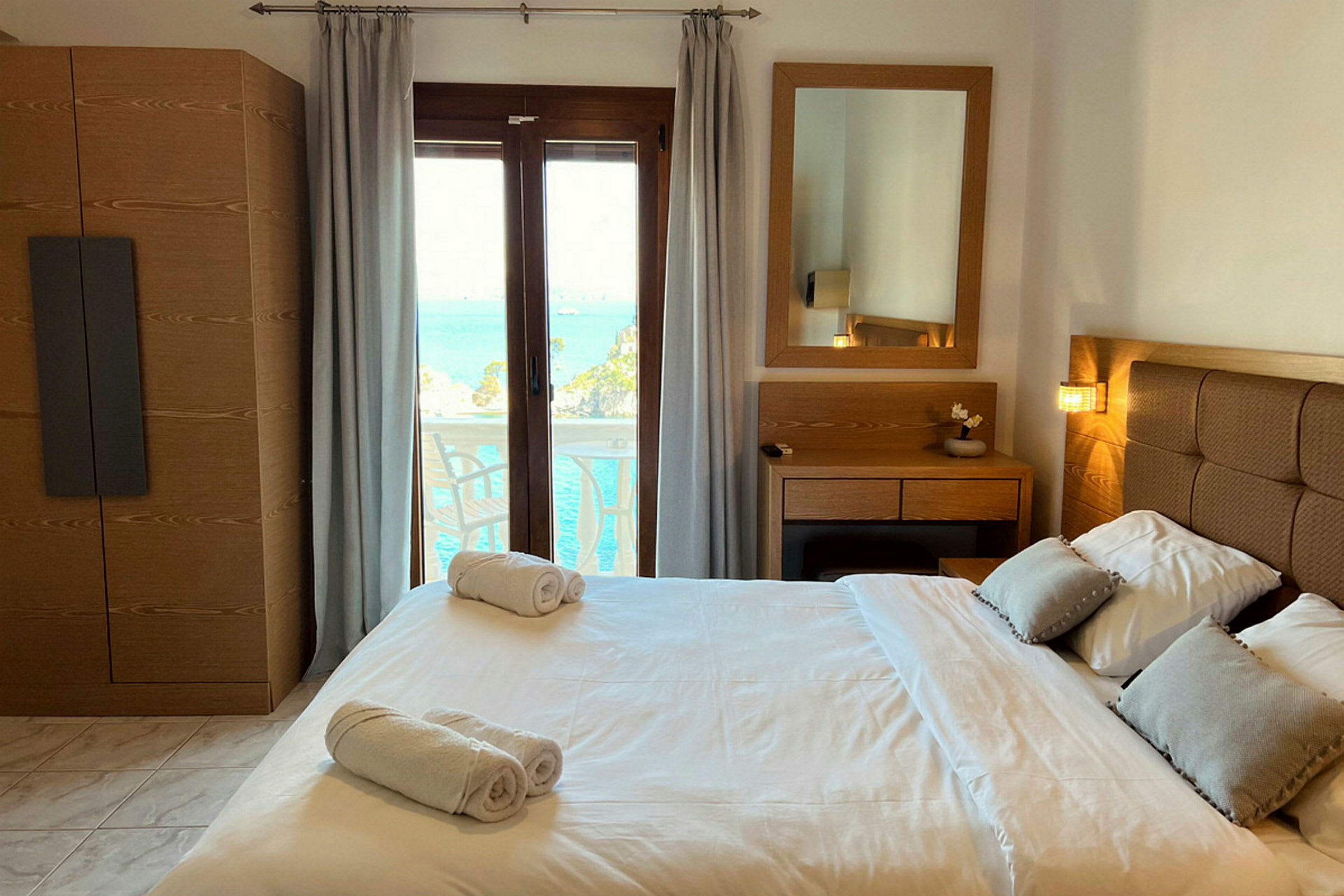 Et hotellrom med en dobbeltseng, nattbord, speil og utsikt over havet gjennom et glassvindu. (AI-generert tekst)