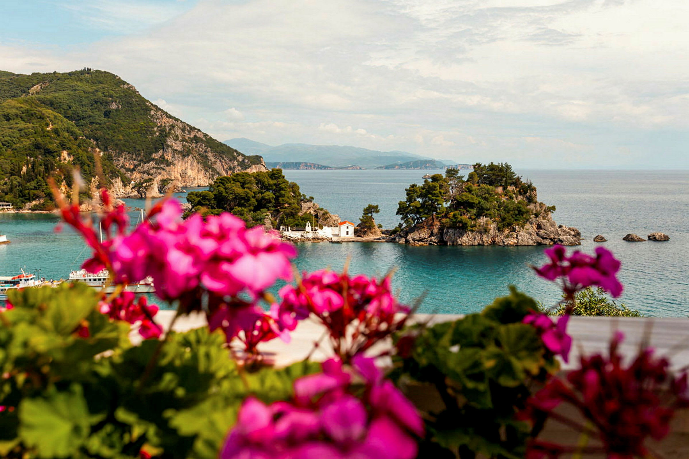 Et naturskjønt landskap med en liten øy i havet, omgitt av grønne trær og fjell i bakgrunnen. Foran er det fargerike blomster. (AI-generert tekst)