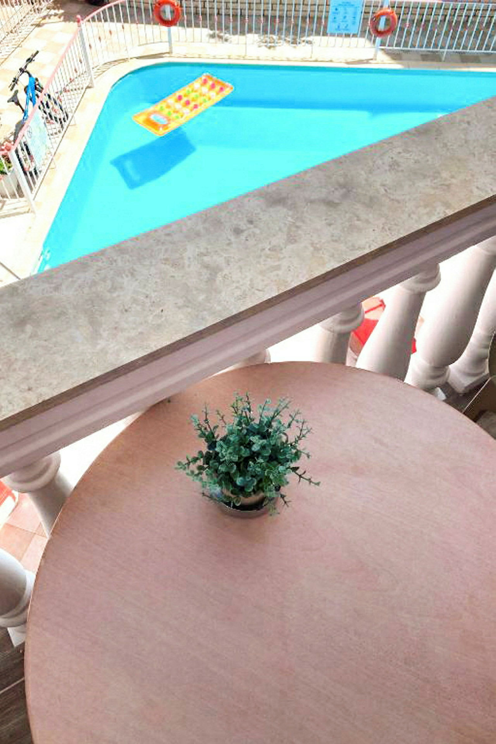 En utsikt fra en balkong mot et basseng med en badeleke og et bord med en potteplante. (AI-generert tekst)