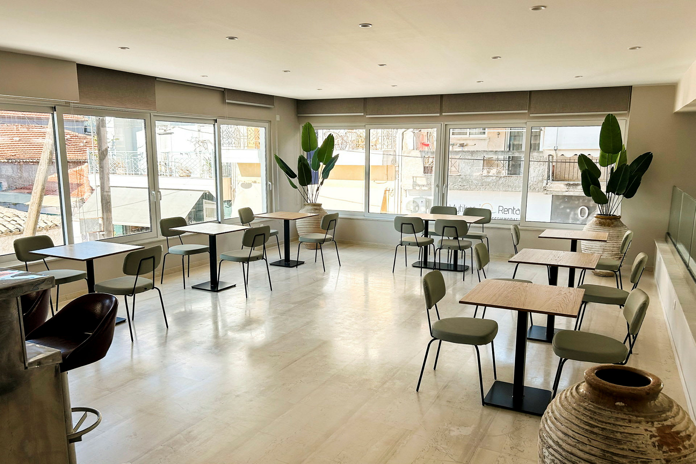 En lys kafé med bord og stoler samt store vinduer som slipper inn naturlig lys. Flere potteplanter pryder rommet. (AI-generert tekst)