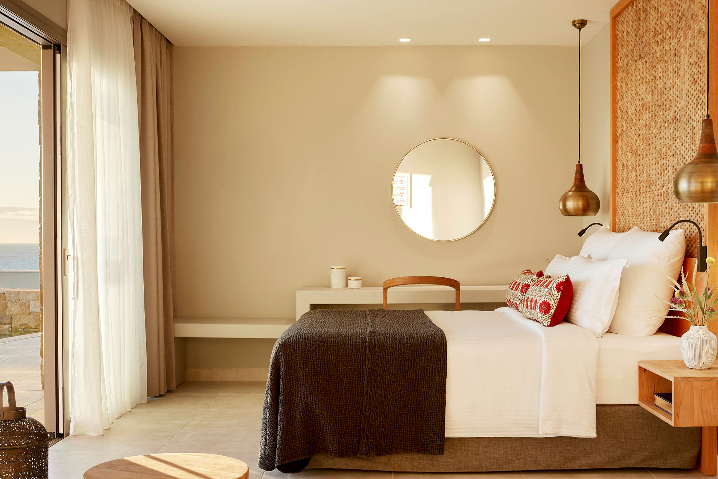 Et hotellrom med en stor seng, runde dekorative lamper og utsikt mot havet gjennom en glassparti. (AI-generert tekst)