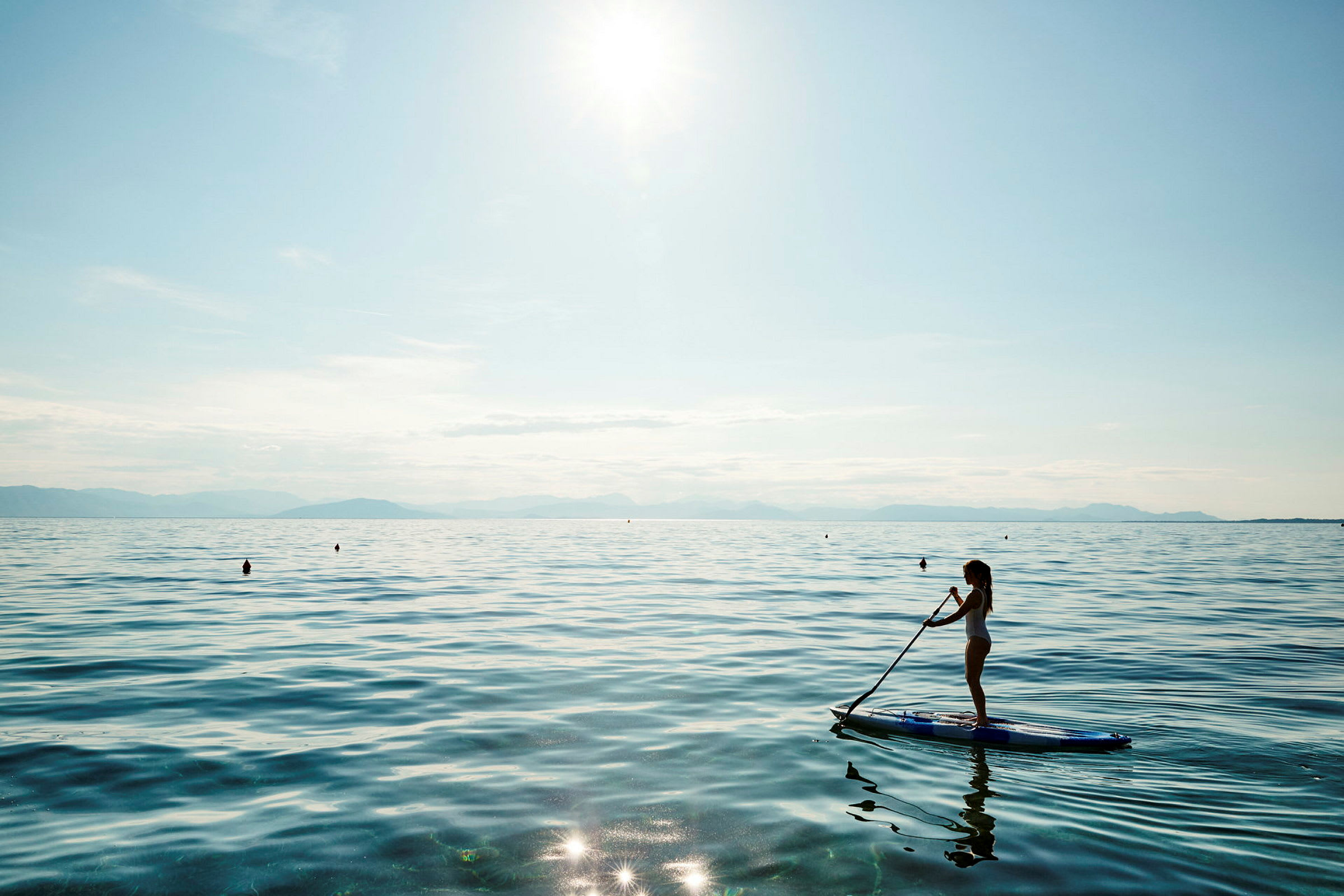 Kvinne som står på et paddleboard på en rolig innsjø med solskinn og fjell i bakgrunnen. (AI-generert tekst)