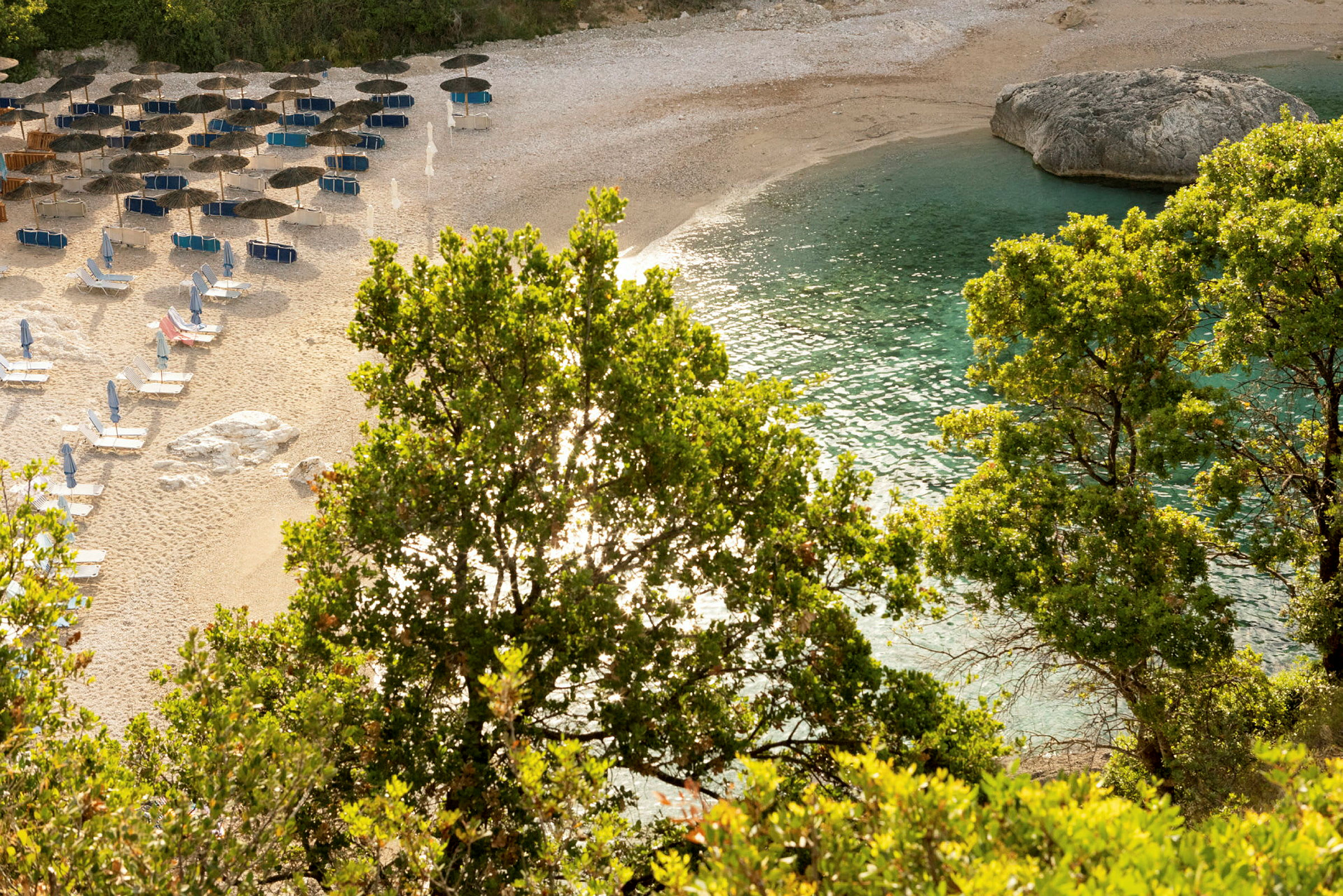 Strand med parasoller, solsenger, frodig vegetasjon og klart blått vann. (AI-generert tekst)