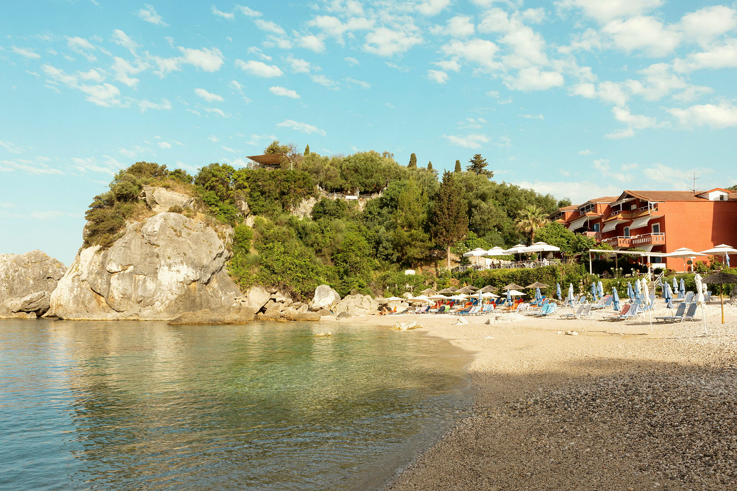 En steinete strand ved klart vann med solstoler og parasoller foran frodig grønt og bygninger. (AI-generert tekst)