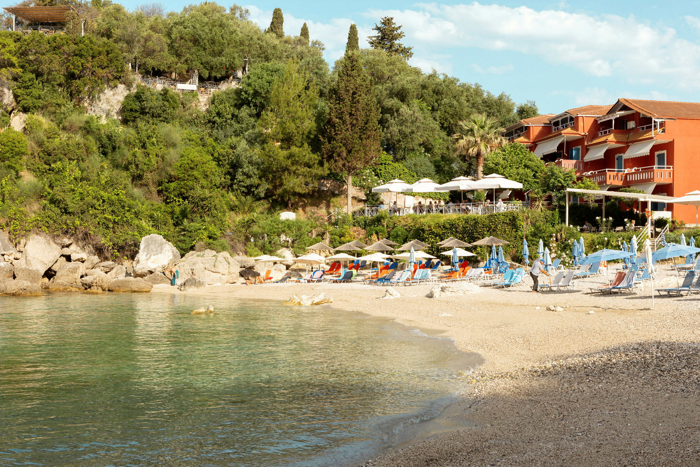 Strand med solstoler og parasoller, frodig skråning og røde bygninger i bakgrunnen. (AI-generert tekst)