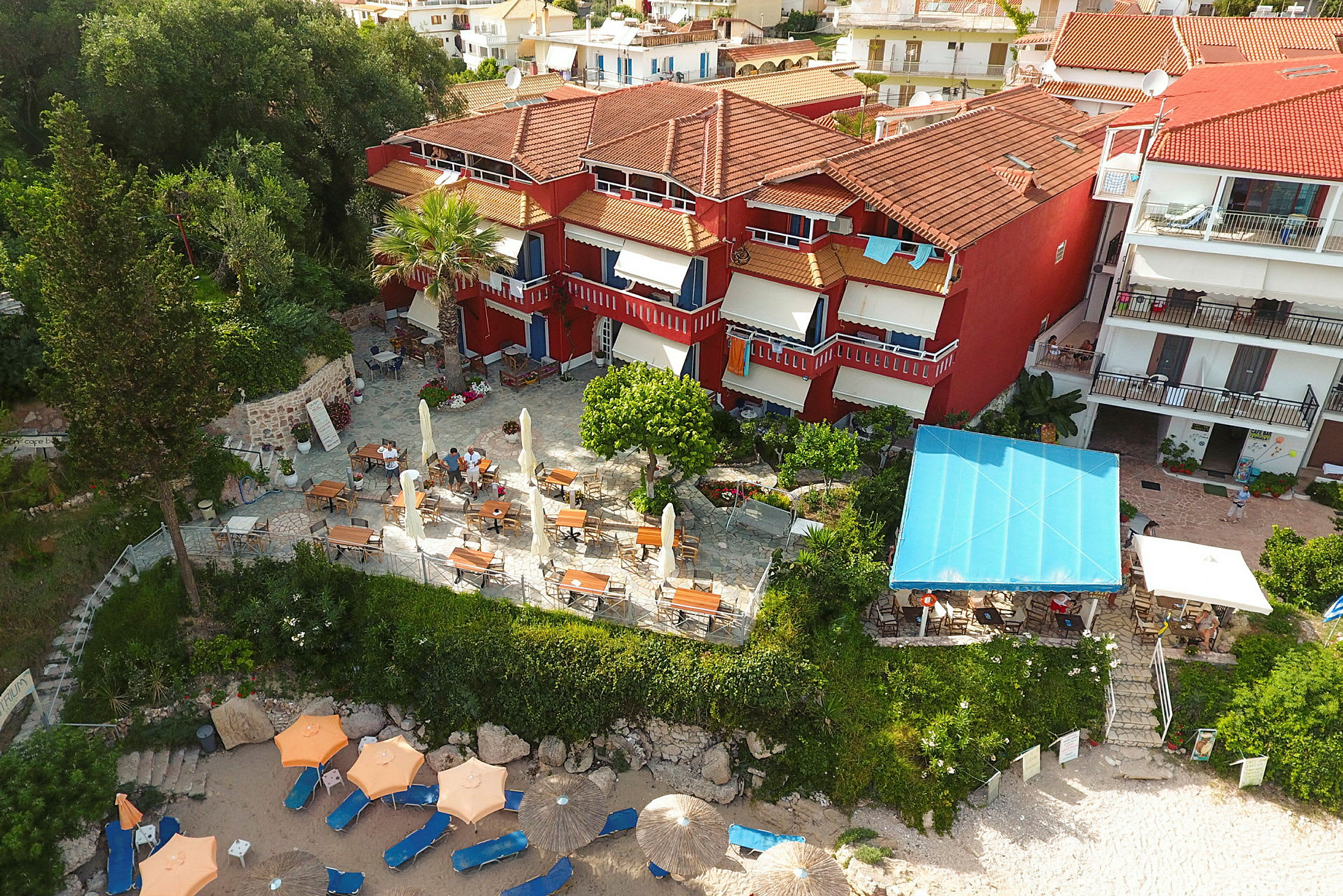 Flyfoto av hotell med røde fasader, en terrasse med bord og stoler samt parasoller på en sandstrand. (AI-generert tekst)