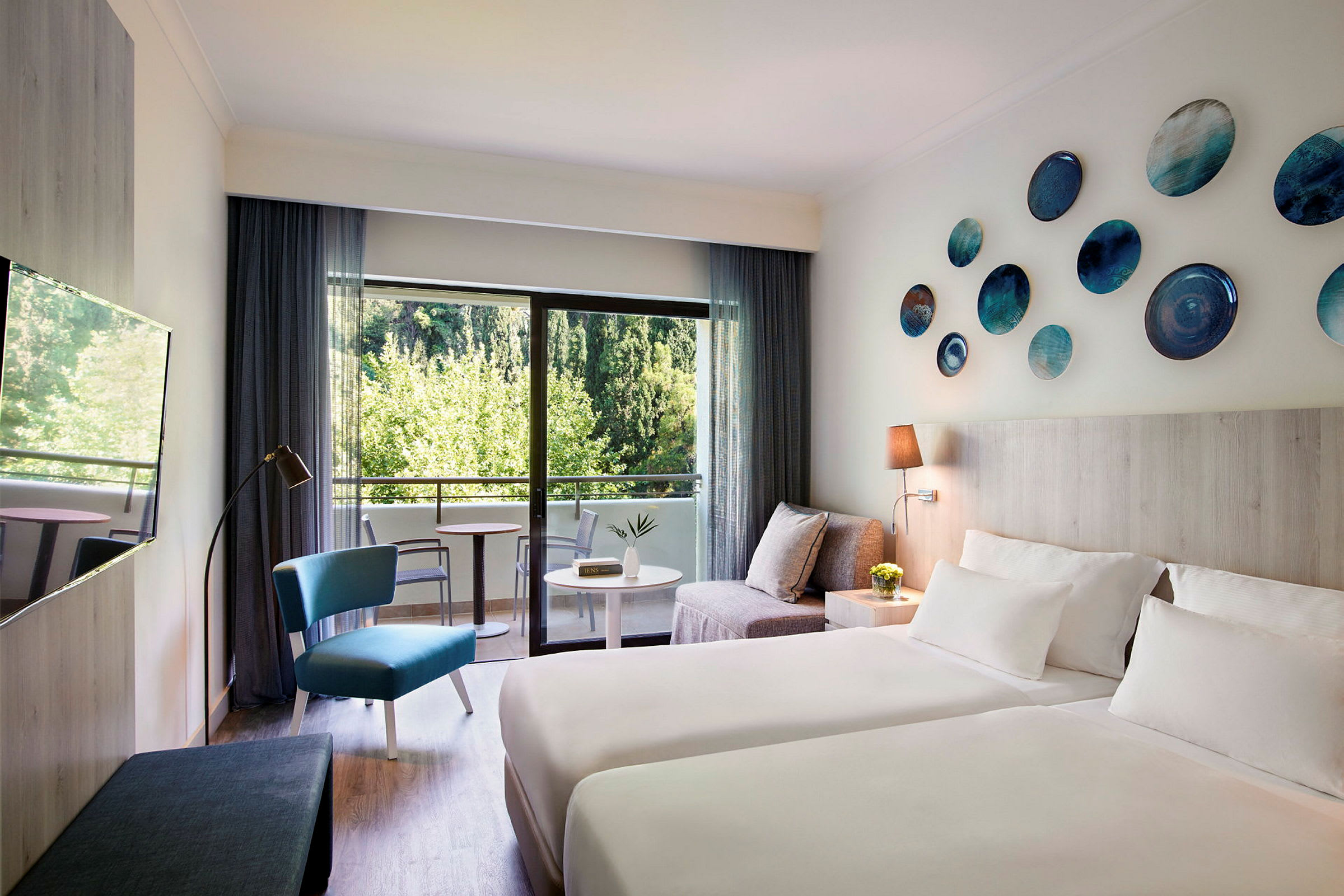 Moderne innredet hotellrom med to senger, en sittegruppe og balkong med utsikt over grøntområder. (AI-generert tekst)
