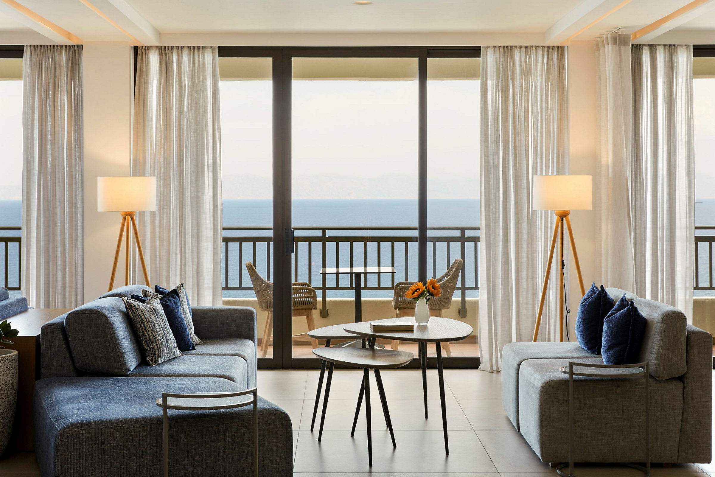 Stue med sofaer og bord, store glassvinduer som vender mot havet. (AI-generert tekst)