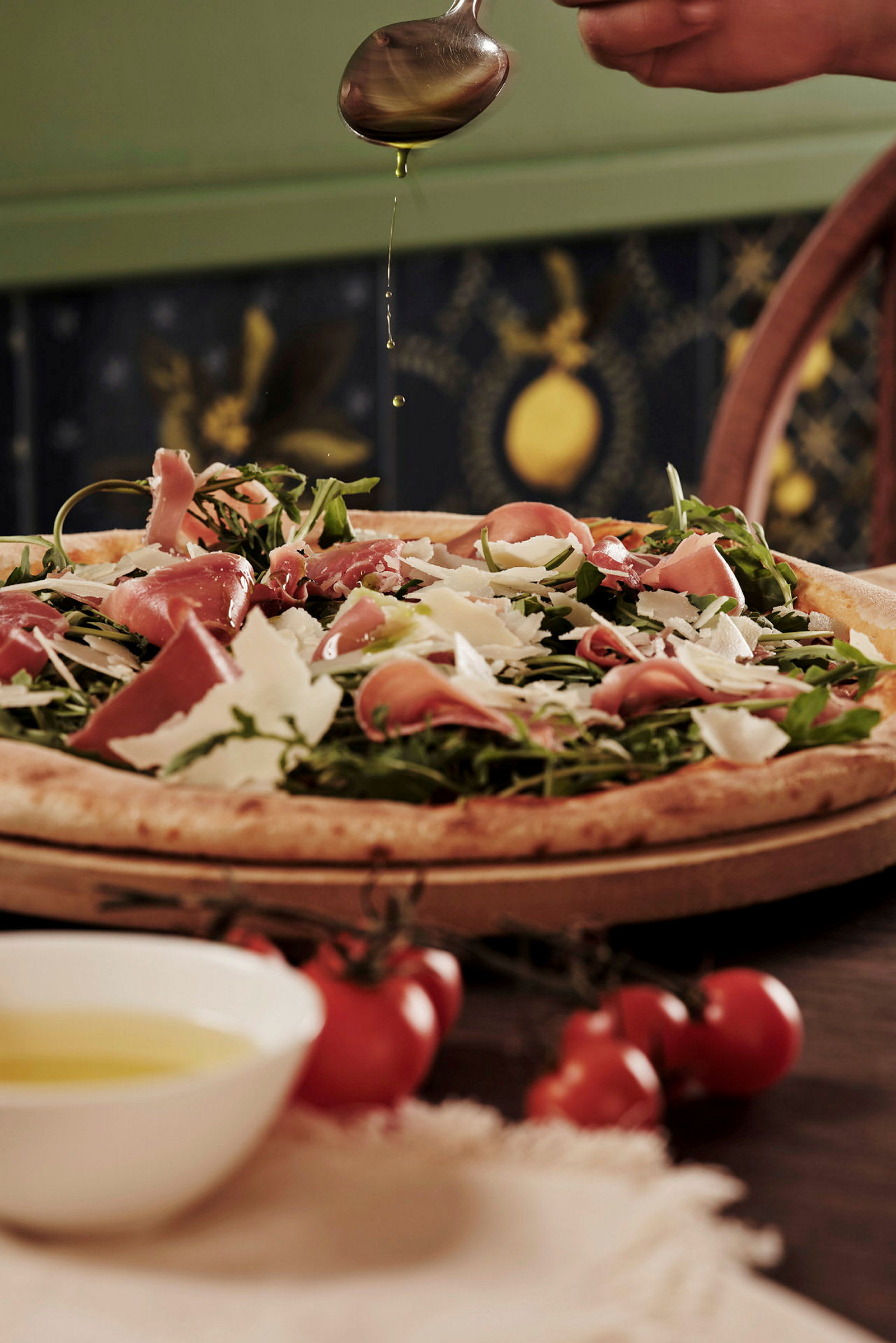 En pizza toppet med skinke, ruccola og parmesan, en hånd drypper olivenolje over den. (AI-generert tekst)