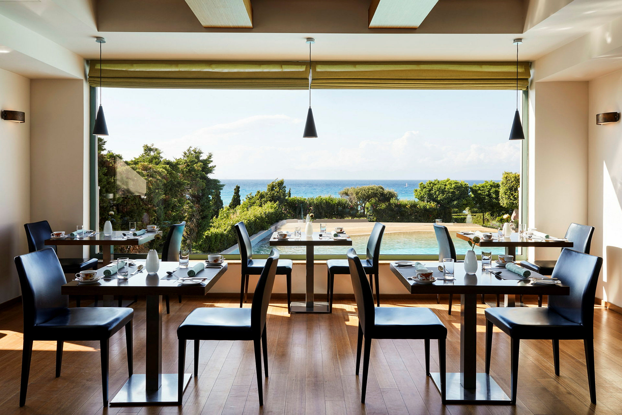 Et lyst restaurantrom med dekkede bord og stoler, med utsikt over et basseng, grønne trær og havet i bakgrunnen. (AI-generert tekst)