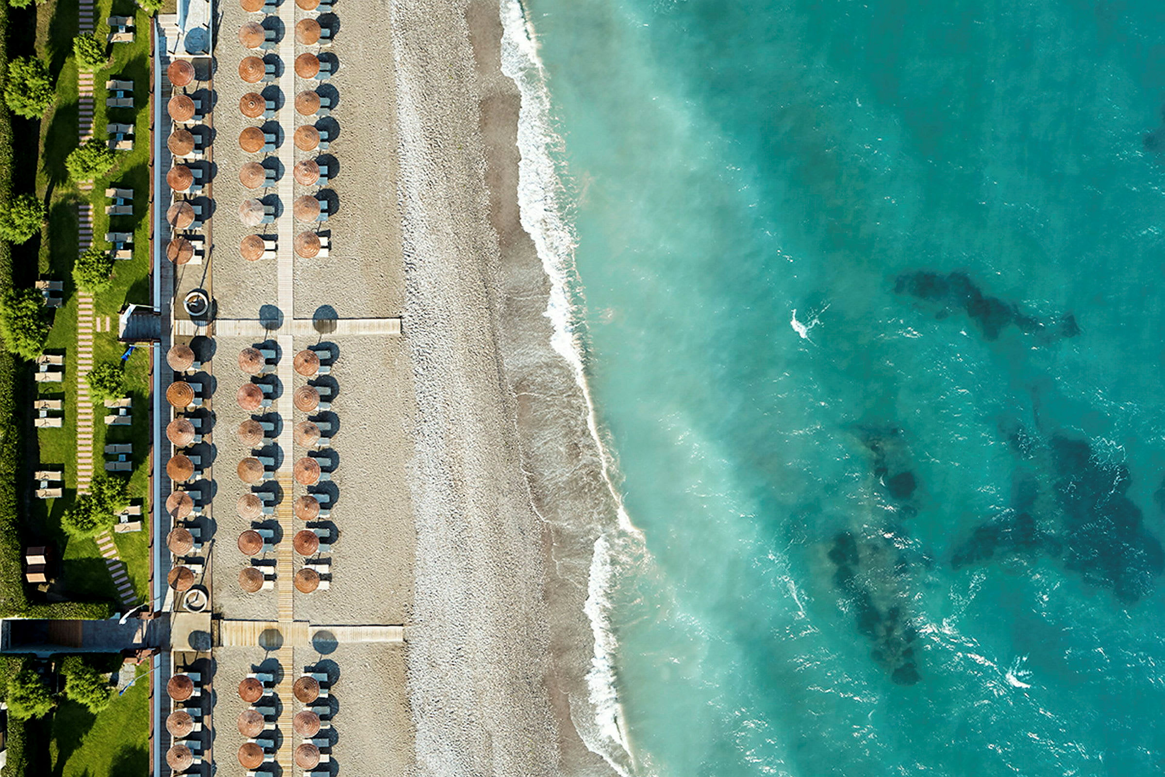 Flyfoto av en strand med parasoller og solsenger ved siden av en turkisblå havstrand. (AI-generert tekst)