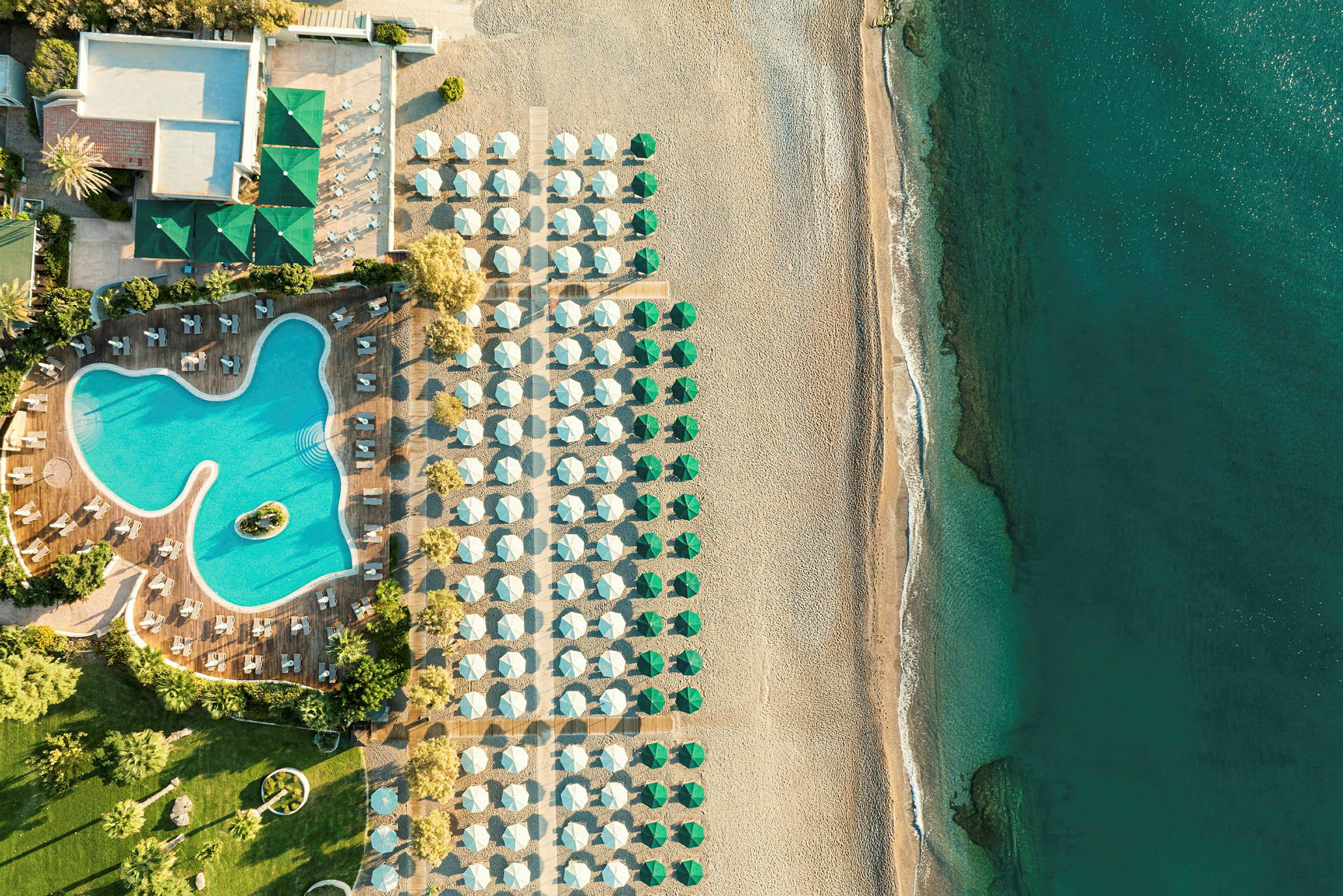 Flyfoto av hotellbasseng med omkringliggende solsenger, ved siden av en strand med parasoller og hav. (AI-generert tekst)
