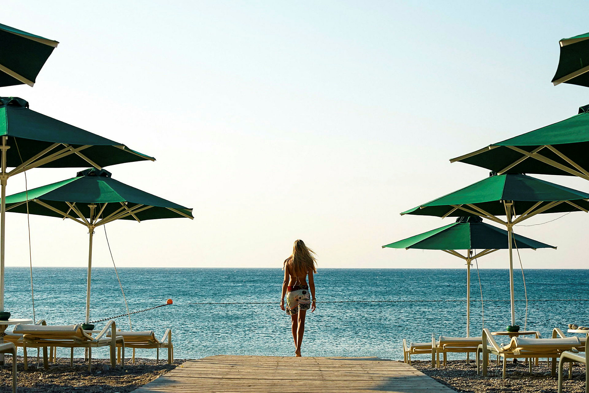 En person går mot havet mellom grønne parasoller og solsenger på stranden. (AI-generert tekst)