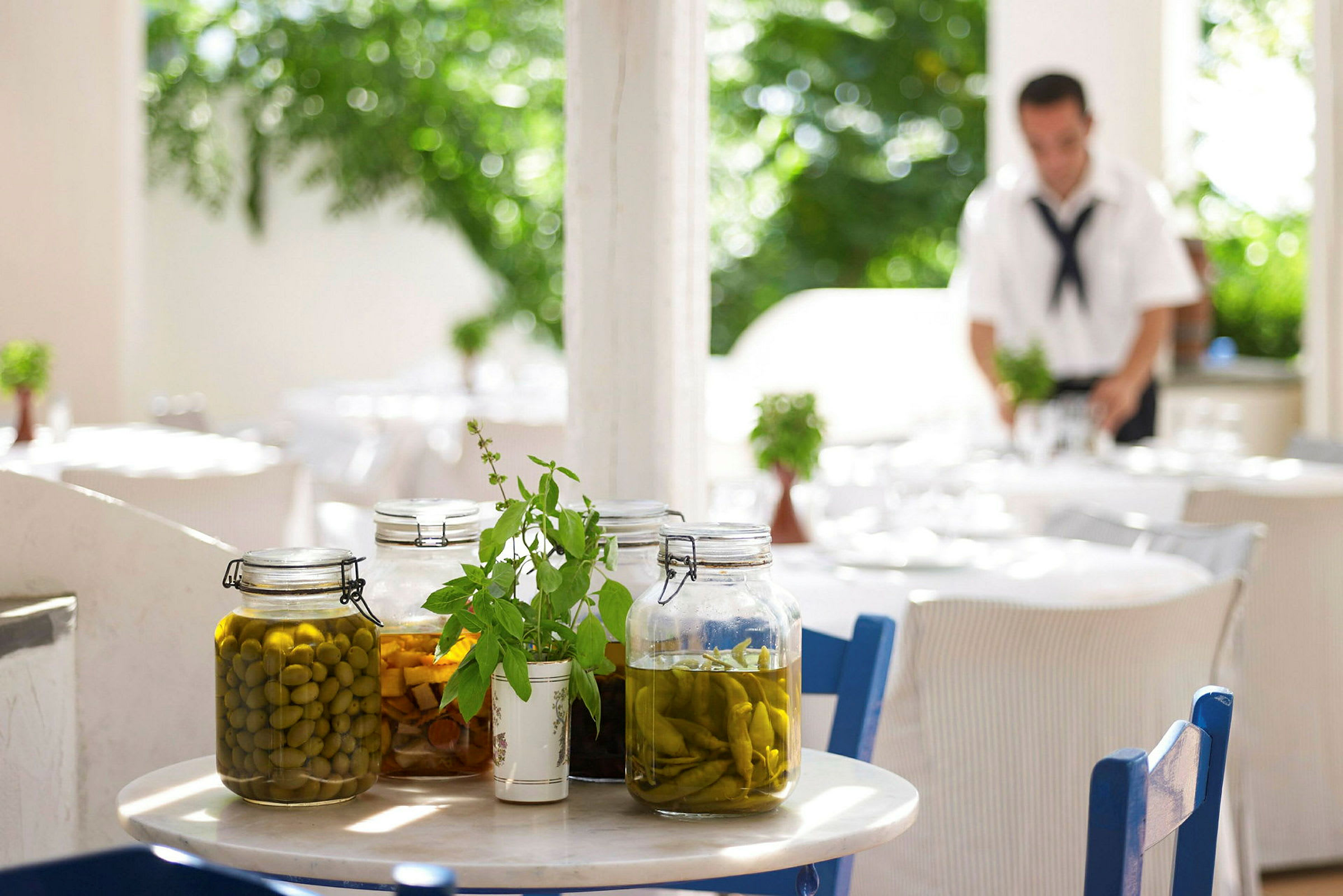 Et bord med glasskrukker fylt med syltede grønnsaker og urtepynt. Bakgrunnen viser en utendørsrestaurant. (AI-generert tekst)